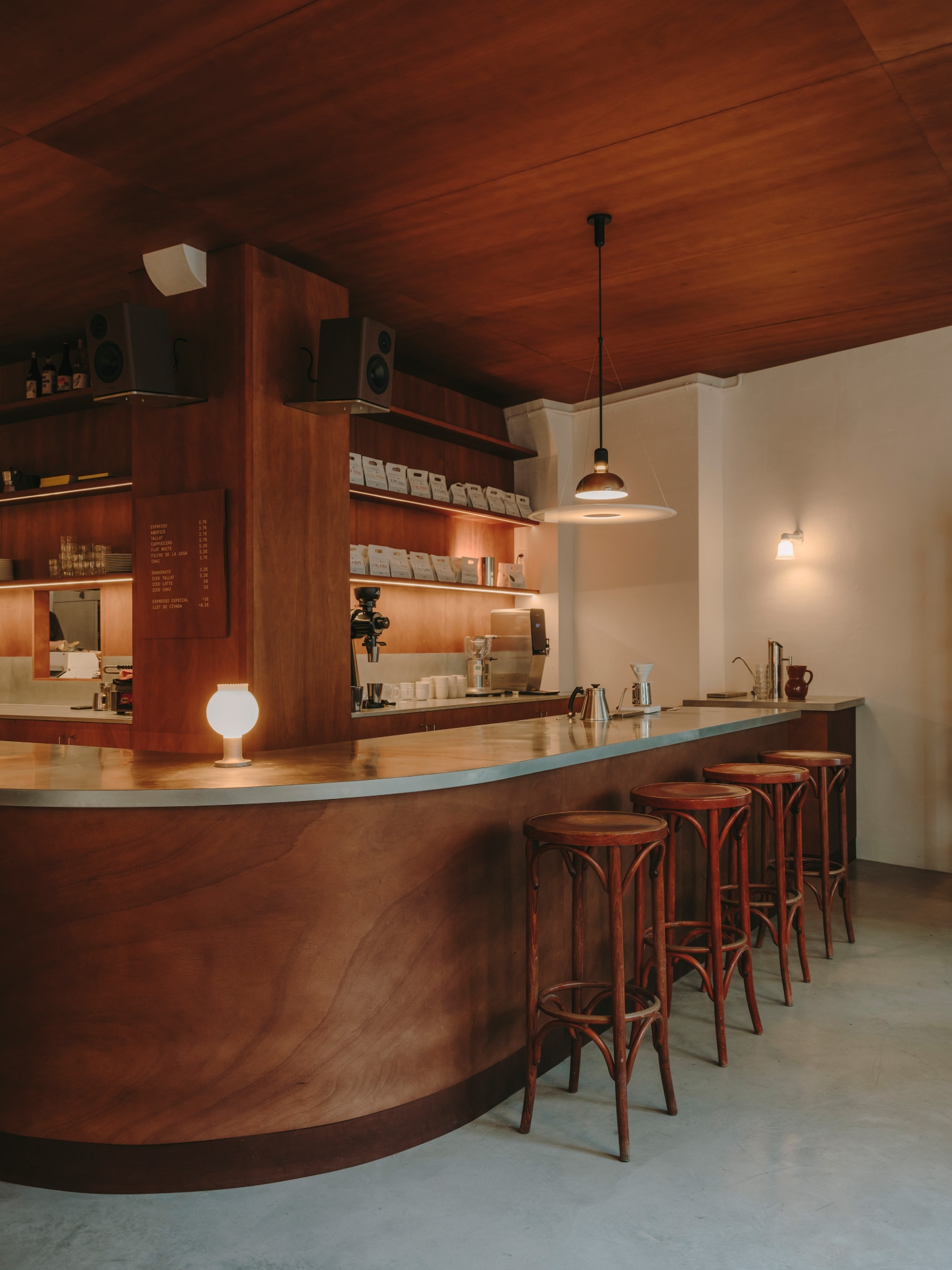 Nomad Coffee Frutas Selectas by Skye Maunsell and Jordi Veciana — The café counter occupies center frame, its curved timber form clad in zinc sheeting and anchored by a globe pendant light integrated into the structure.