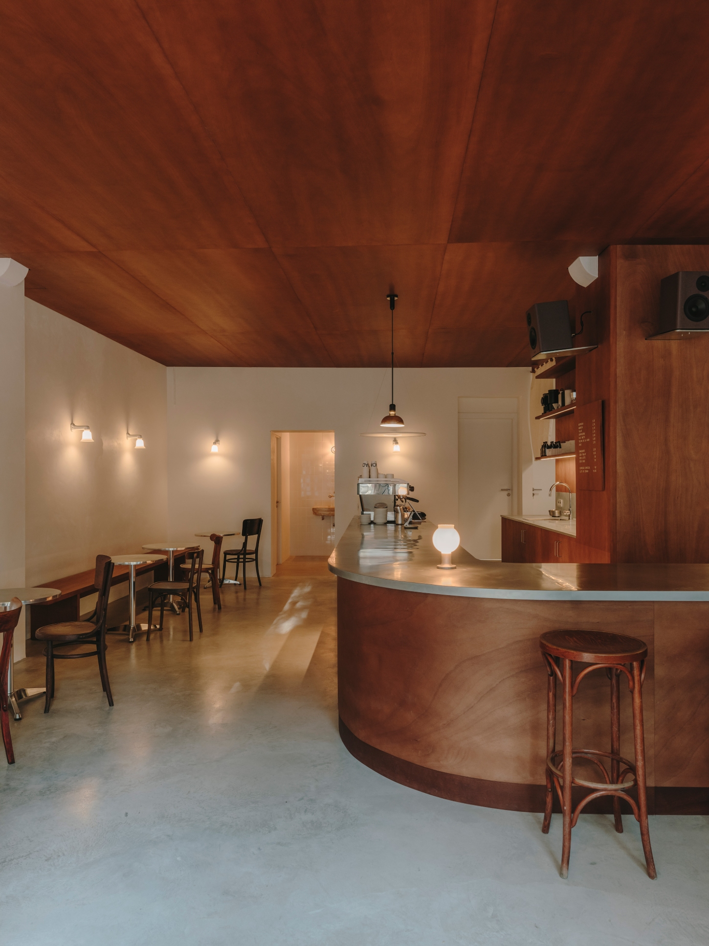 Nomad Coffee Frutas Selectas by Skye Maunsell and Jordi Veciana — The curved timber bar counter dominates the foreground, its walnut stain darkened to near-burgundy, topped with a zinc sheet surface that catches pendant light.