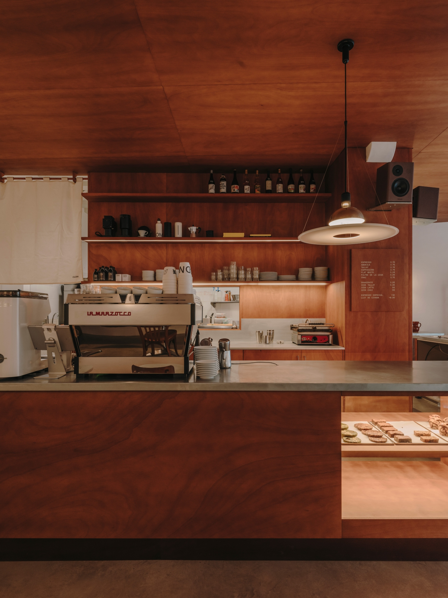 Nomad Coffee Frutas Selectas by Skye Maunsell and Jordi Veciana — The espresso machine anchors the counter's left side, positioned adjacent to a white refrigerator and framed by open shelving displaying bottles, equipment,...