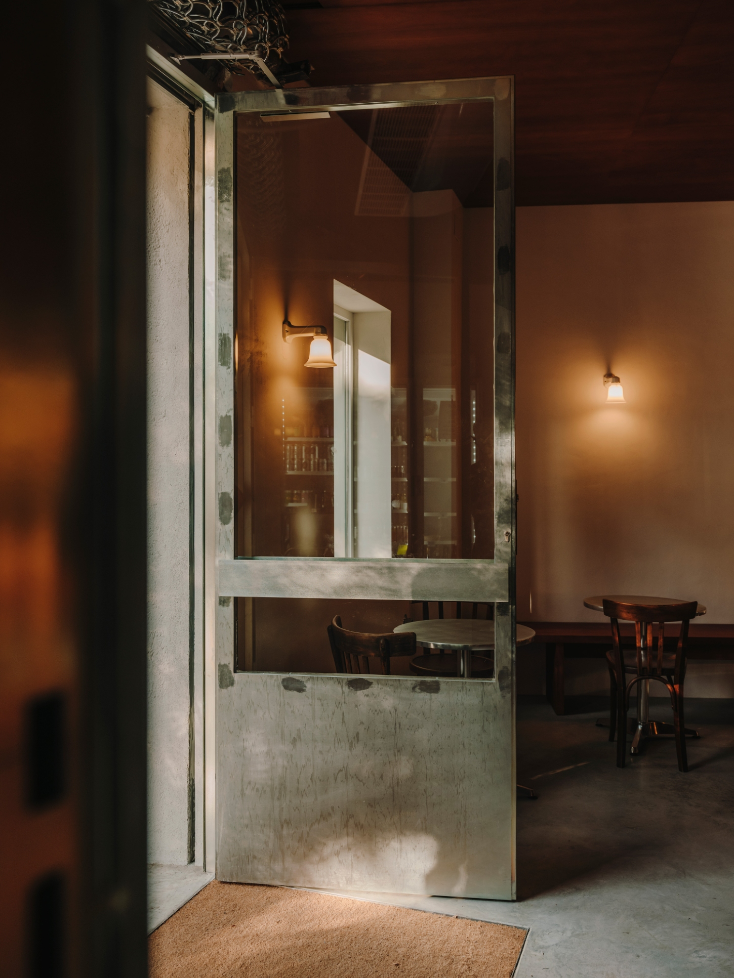 Nomad Coffee Frutas Selectas by Skye Maunsell and Jordi Veciana — A threshold view into Nomad Coffee's dining zone, framed through a weathered metal-framed glass door with zinc sheet lower panel.