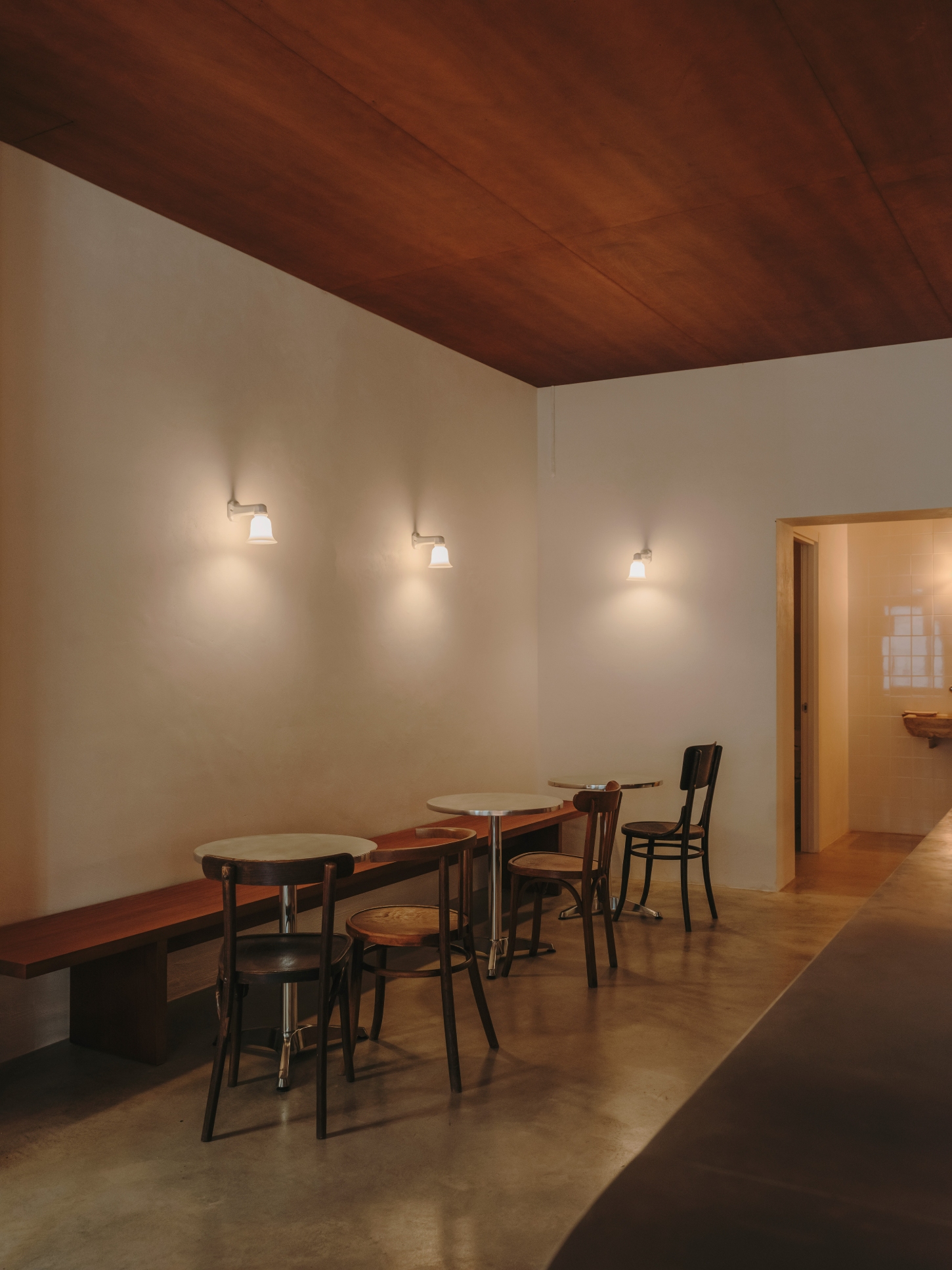 Nomad Coffee Frutas Selectas by Skye Maunsell and Jordi Veciana — The dining zone features a linear arrangement of small tables with bentwood café chairs positioned against cream-rendered walls beneath wall-mounted pendant...