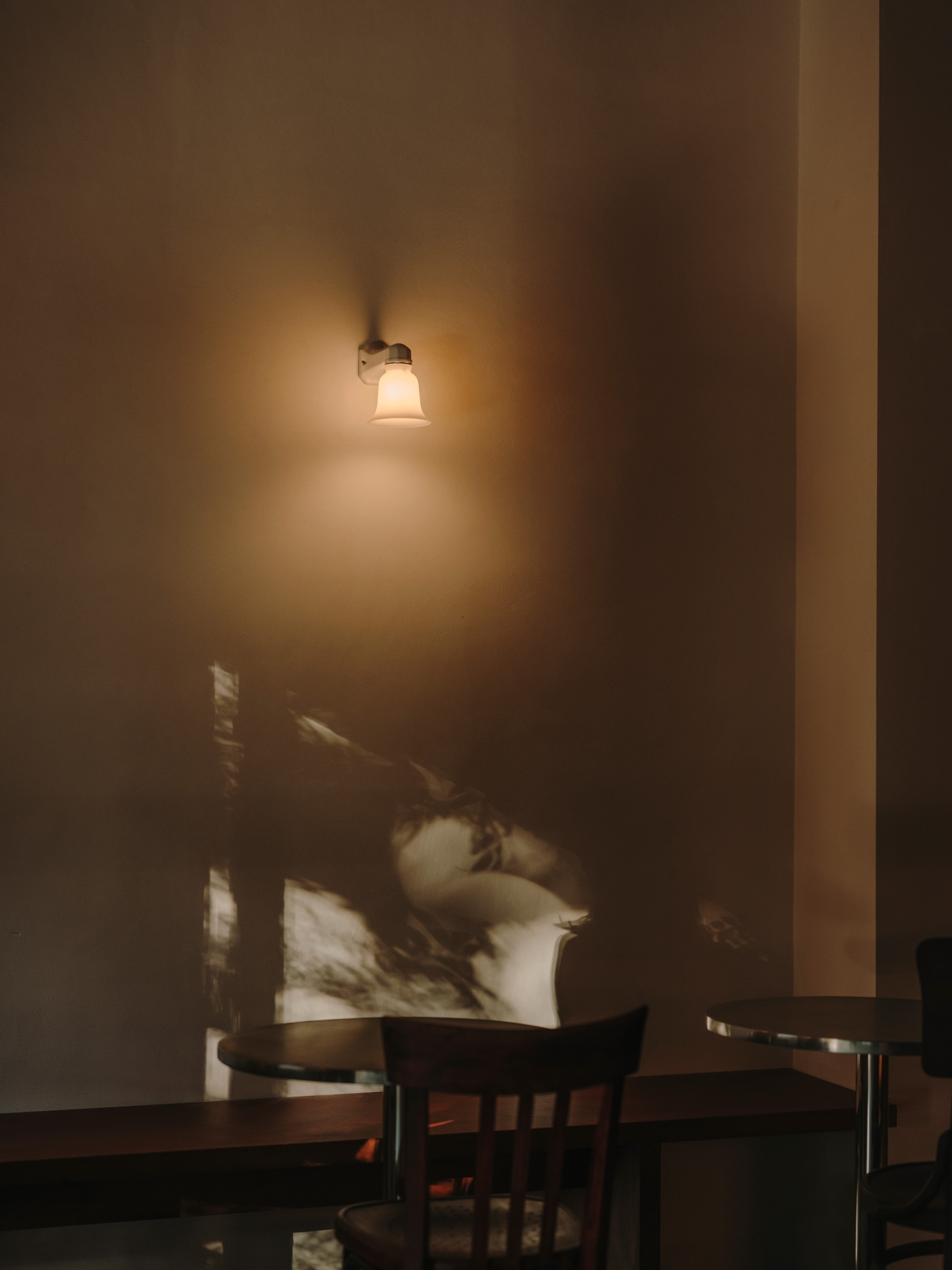 Nomad Coffee Frutas Selectas by Skye Maunsell and Jordi Veciana — A pendant light suspended from the dark-stained timber ceiling casts a concentrated pool of warm light onto the lime mortar wall, creating pronounced shadow...