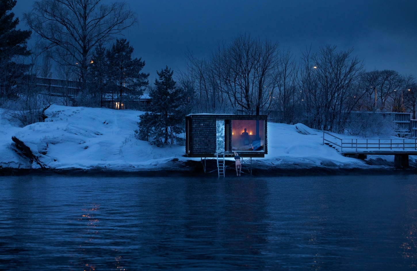 The Hotspot Oslo Works — A timber sauna elevated on wooden stilts extends into dark water, its burnt-wood shingle exterior framing a single rectangular aperture that glows with...