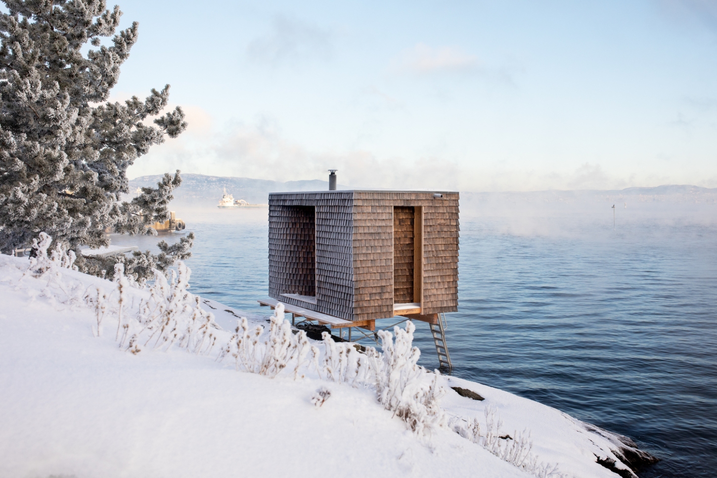 The Hotspot Oslo Works — A timber-clad sauna structure elevated on wooden stilts juts into calm water on the Nesodden Peninsula.
