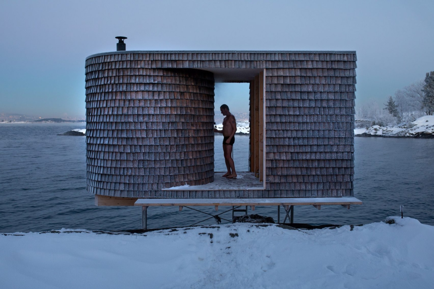The Hotspot Oslo Works — A wooden sauna structure elevated on stilts extends into a frozen waterscape on Norway's Nesodden Peninsula.