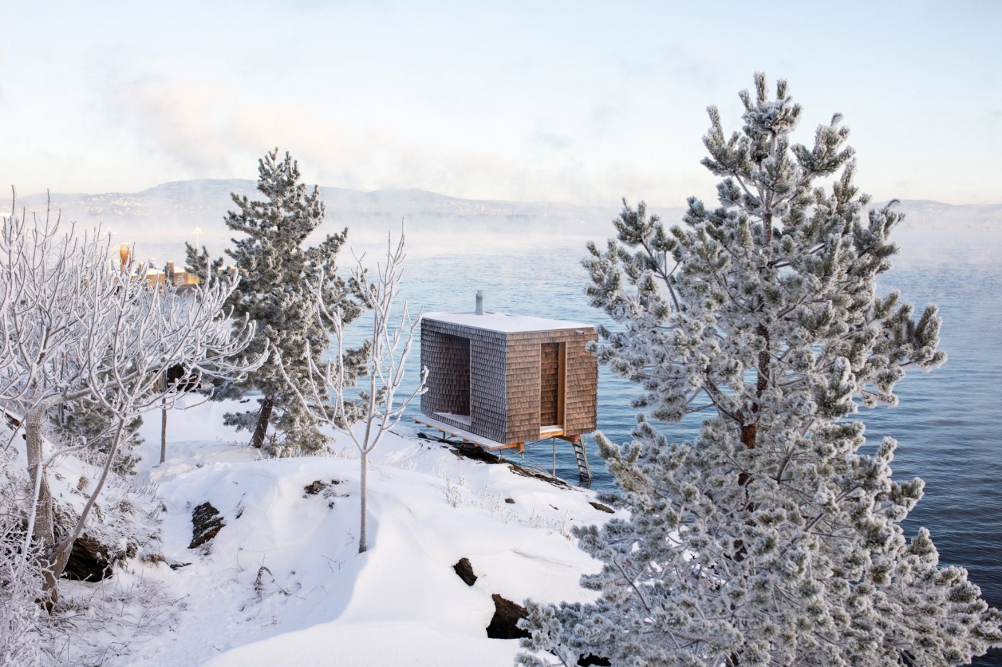 The Hotspot Oslo Works — A timber sauna elevated on wooden stilts extends into the frozen shoreline of the Nesodden Peninsula, its dark cedar-shingle exterior punctuated by a single...