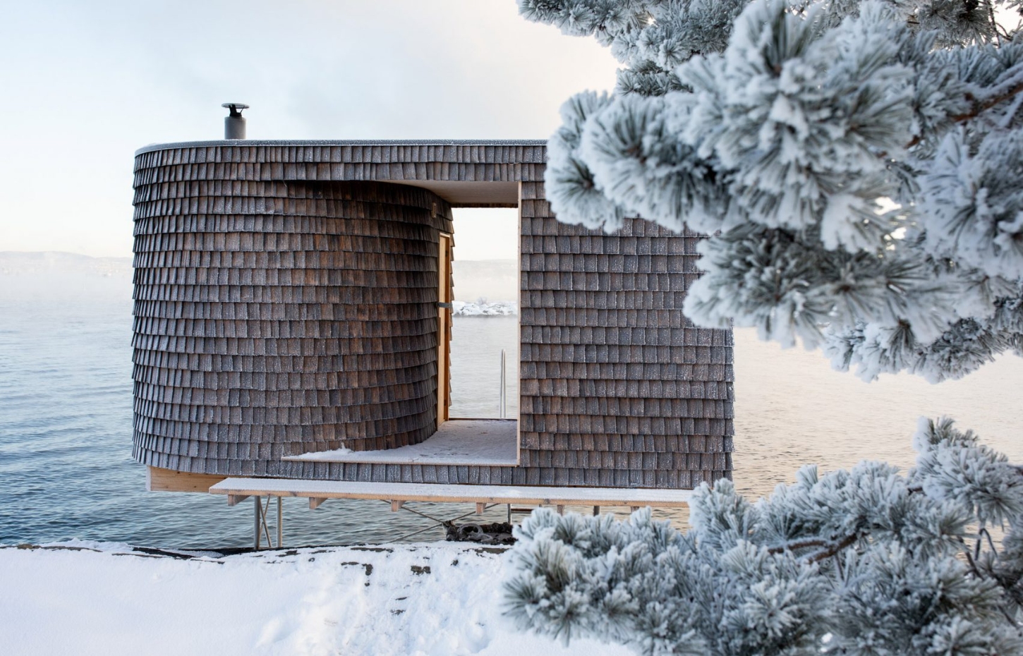 The Hotspot Oslo Works — A wood-shingled sauna structure sits elevated on wooden stilts above the waterline on the Nesodden Peninsula.