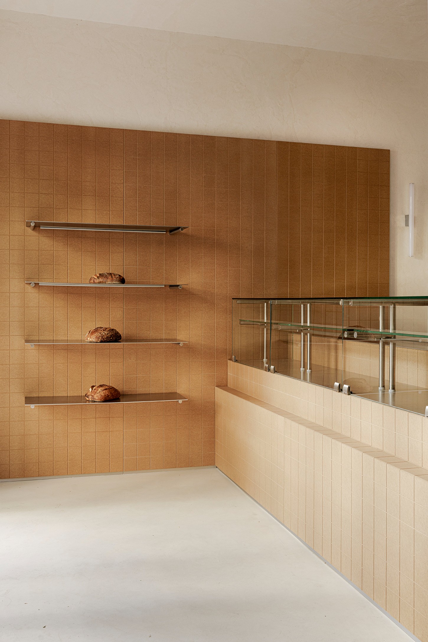 Madreamiga Madrid by estudio DIIR — A bakery display nook features a gridded terracotta tile wall with four horizontal stainless steel shelves holding artisanal bread loaves in kraft paper.
