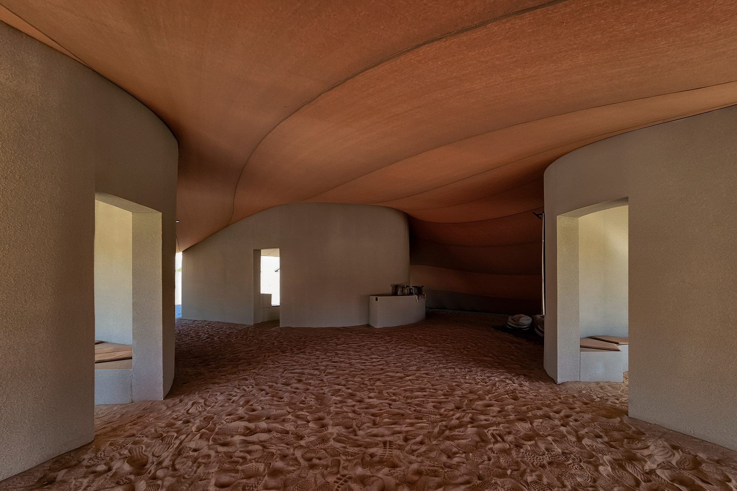 Hegra Viewpoints AlUla desert by Clover Studio — Interior view of a sand-floored pavilion with a billowing terracotta fabric canopy suspended overhead, creating organic undulating forms.