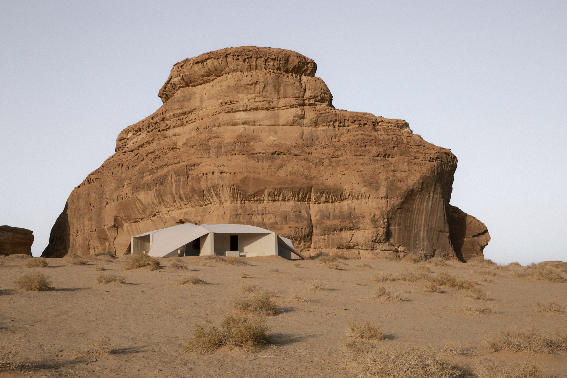 Hegra Viewpoints AlUla desert by Clover Studio — A white fabric canopy billows across the entrance of a contemporary structure carved into a massive sandstone rock formation.