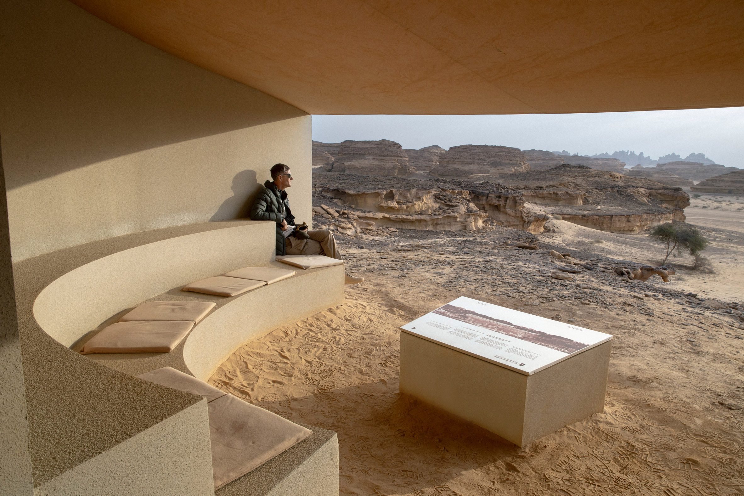 Hegra Viewpoints AlUla desert by Clover Studio — A curved concrete viewing platform anchors a solitary visitor within a monumental desert amphitheater.