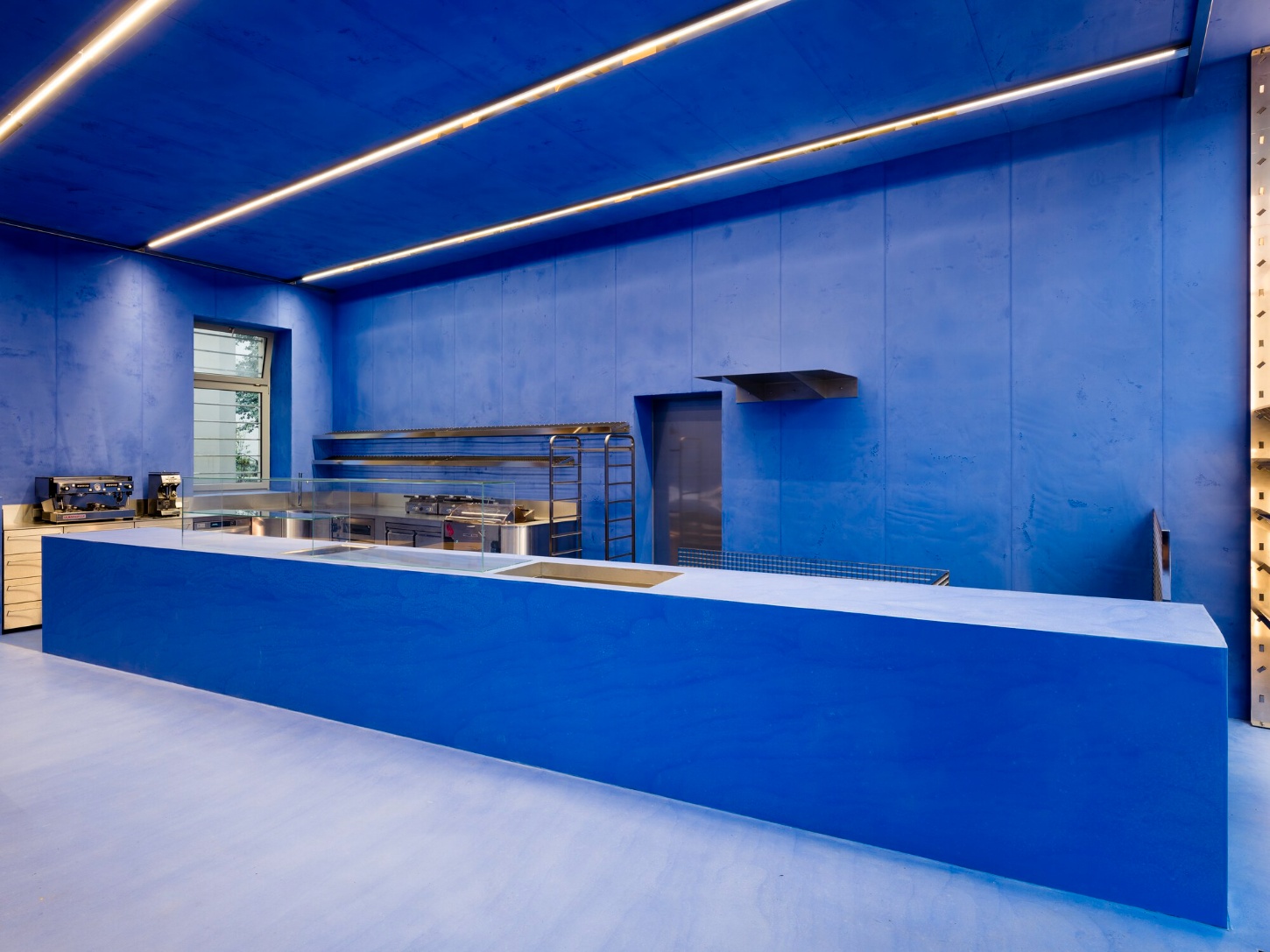 Aera Bakery by Gonzalez Haase AAS — Aera Bakery's service counter extends 13 meters in an unbroken diagonal across the 70m² space, its white laminate top slicing through a monochromatic blue...
