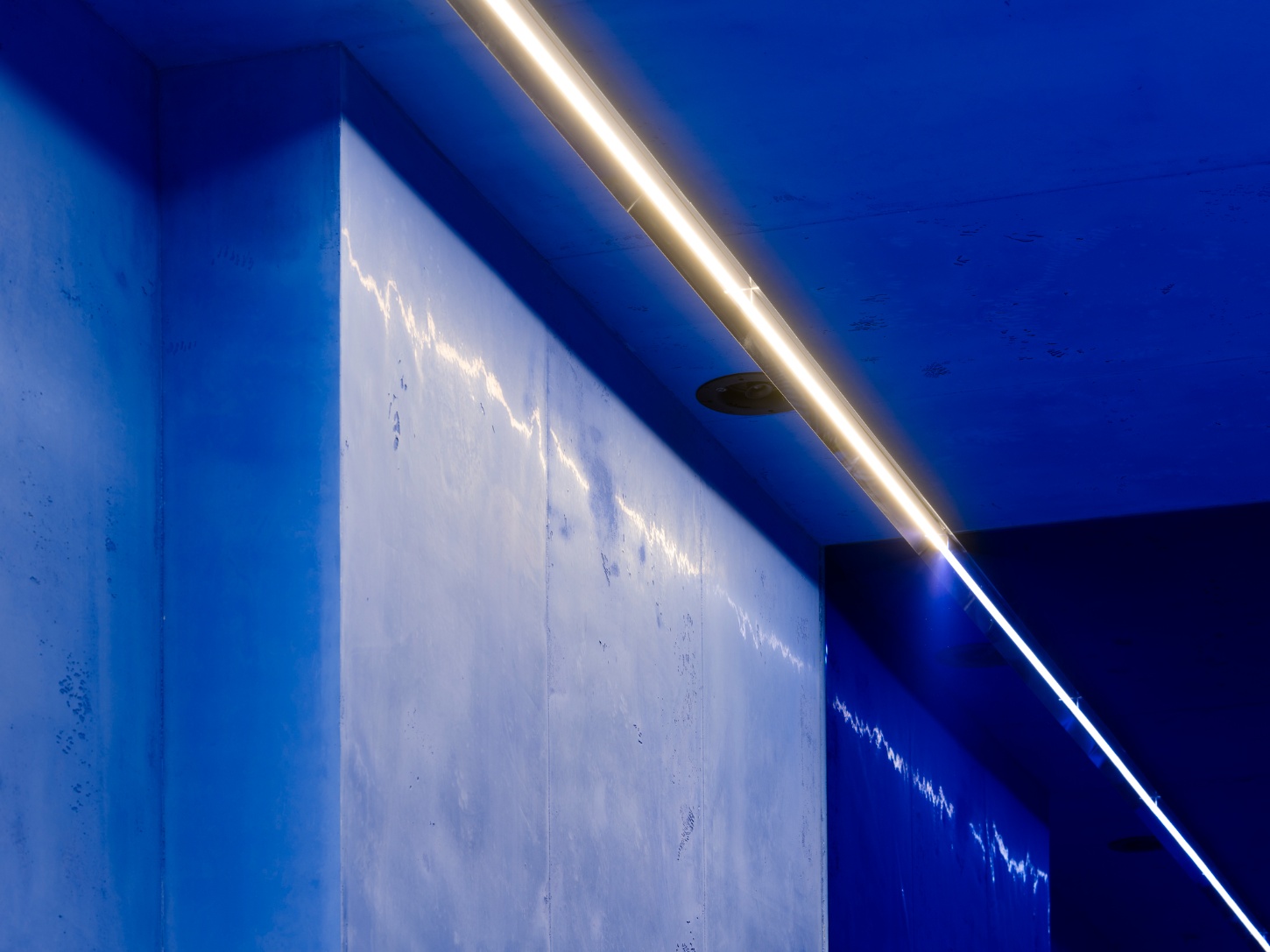 Aera Bakery by Gonzalez Haase AAS — A diagonal linear LED strip fixture traces the ceiling edge in warm white light, creating a sharp contrast against the monochromatic lapis lazuli blue that...