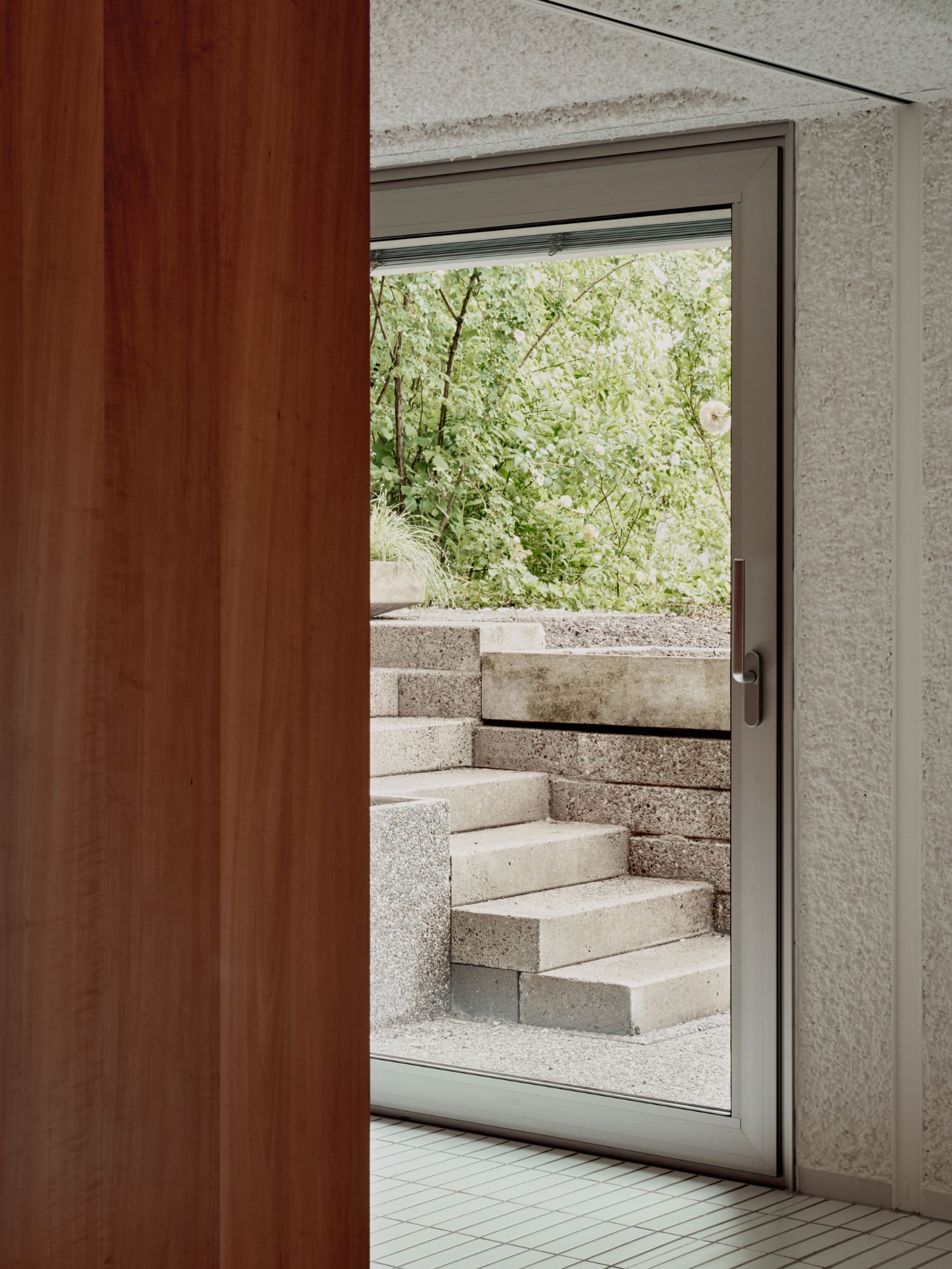 1930’s House Zurich by Ductus by Ductus St — A threshold moment captured through a sliding glass door: concrete steps descend in monolithic blocks, their raw texture catching diffused light.