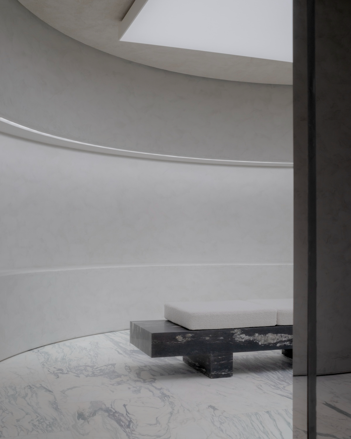 Saint Laurent Flagship in Paris by Sain Laurent — A curved bench with white cushioning sits atop a black-lacquered steel base within a cylindrical white interior.
