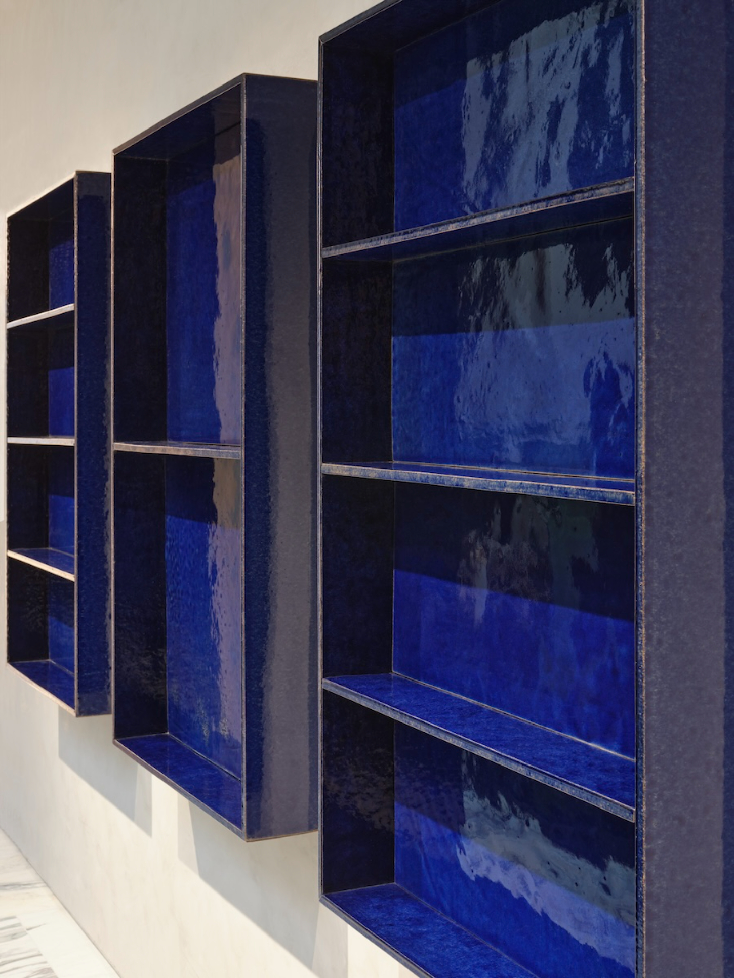 Saint Laurent Flagship in Paris by Sain Laurent — Three modular display units in oxidized steel with deep blue lacquered backing panels, arranged in receding perspective against a white wall.