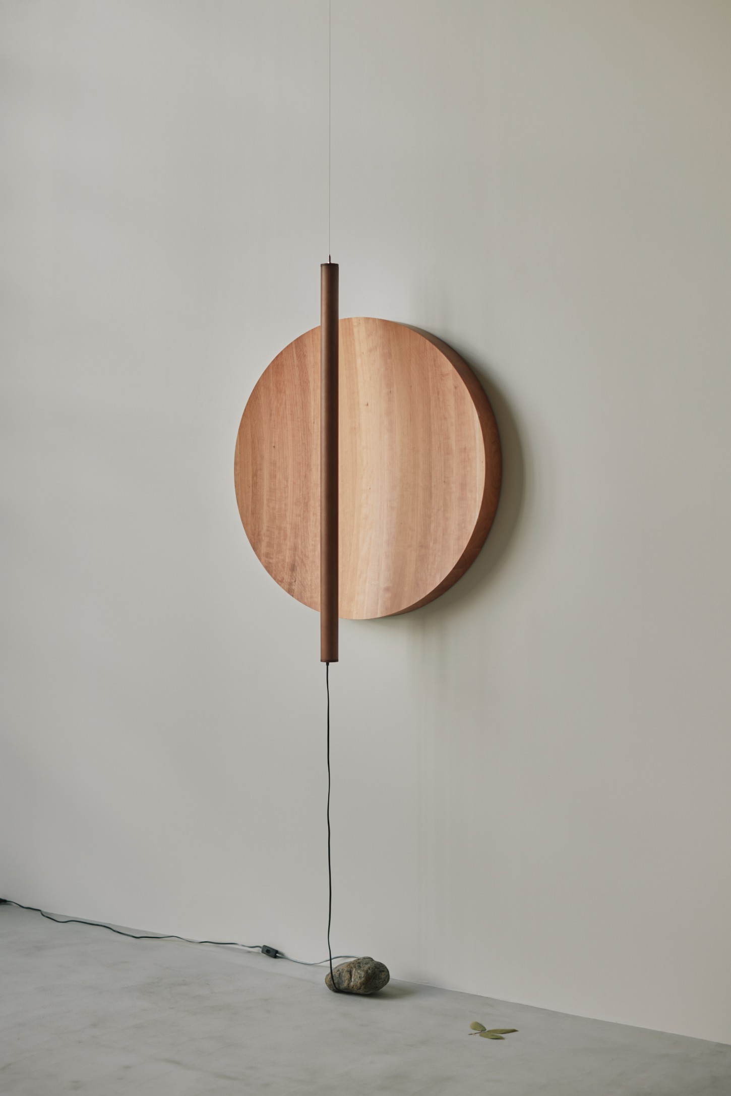 'The Age of Wood' by CHRISTIAN + JADE in Karimoku Research — A suspended wooden disc in contrasting light and dark timber tones is bisected by a vertical dark timber rod extending downward.