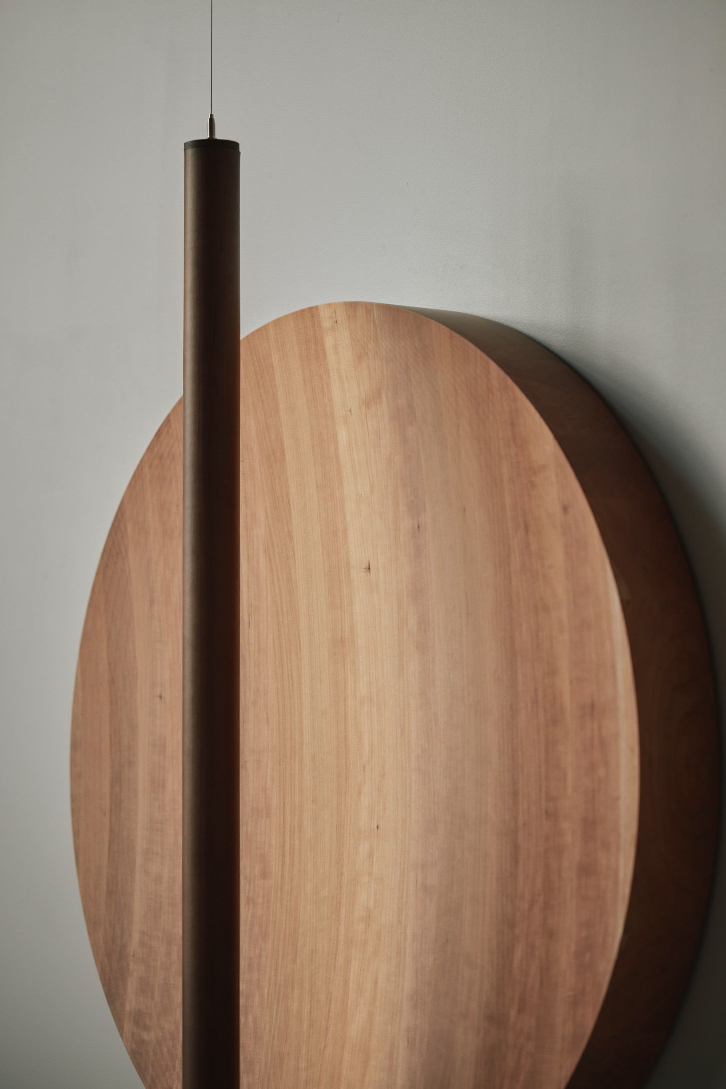 'The Age of Wood' by CHRISTIAN + JADE in Karimoku Research — A cylindrical timber pendant light suspended before a large oval timber panel, part of the modular grid-wall system at Karimoku Research Center.