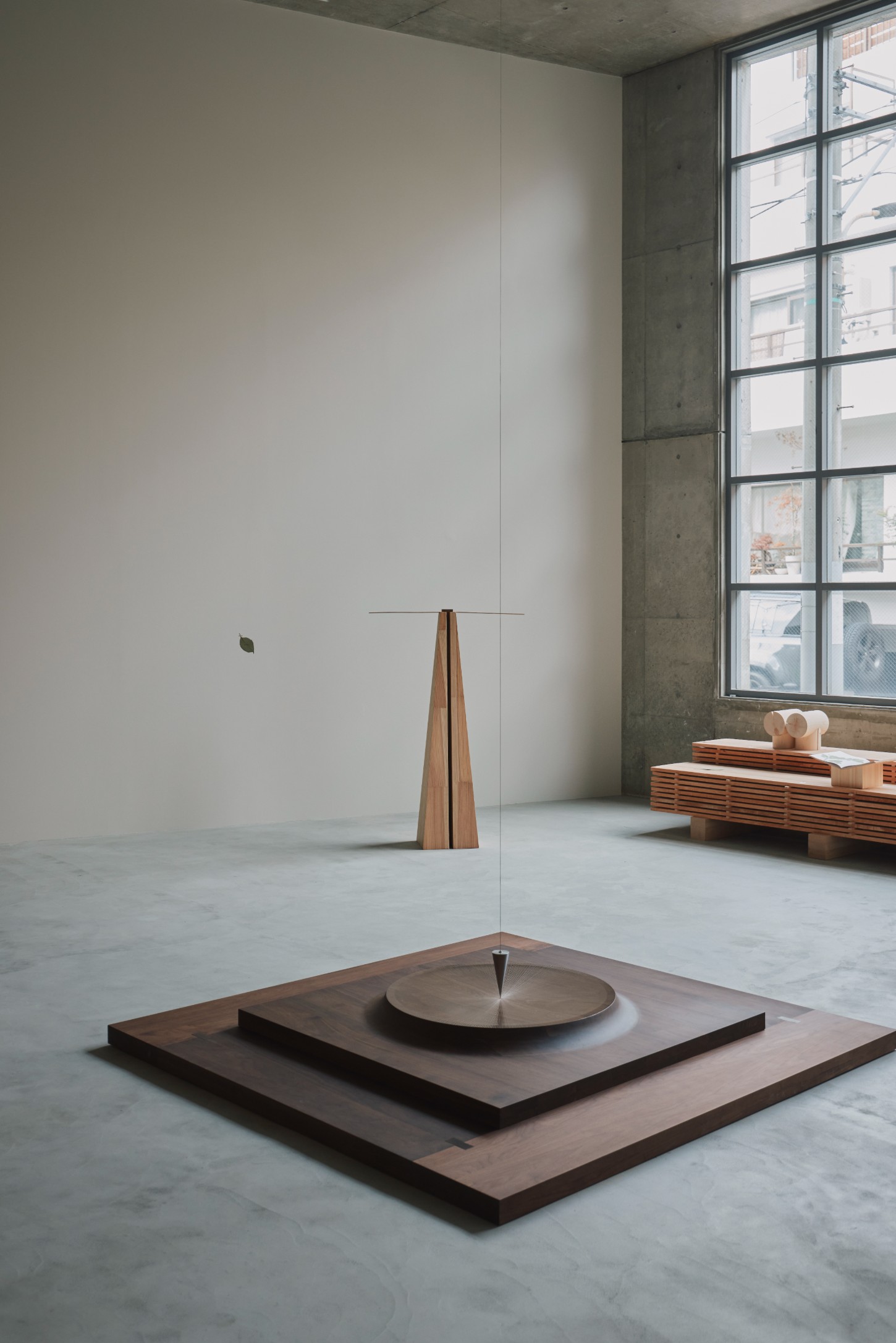 'The Age of Wood' by CHRISTIAN + JADE in Karimoku Research — A timber construction occupies the center of a polished concrete floor: nested square platforms in dark wood rise to support a circular disc with a centered...
