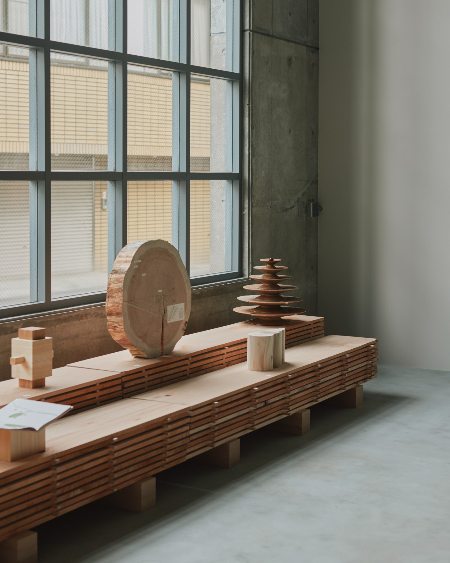 'The Age of Wood' by CHRISTIAN + JADE in Karimoku Research — Within the Karimoku Research Center, a timber bench with horizontal slatted construction supports a curated arrangement of wooden objects.