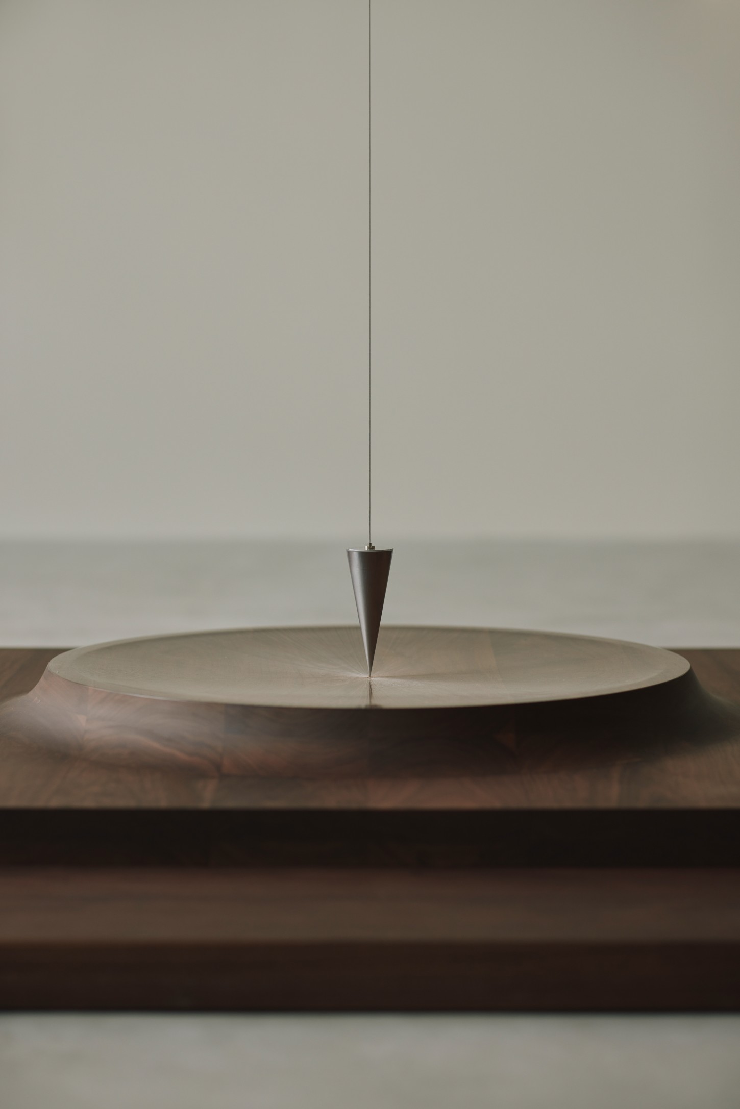 'The Age of Wood' by CHRISTIAN + JADE in Karimoku Research — A suspended metal cone hangs above a circular timber platform, its pointed tip nearly touching the pale surface below.