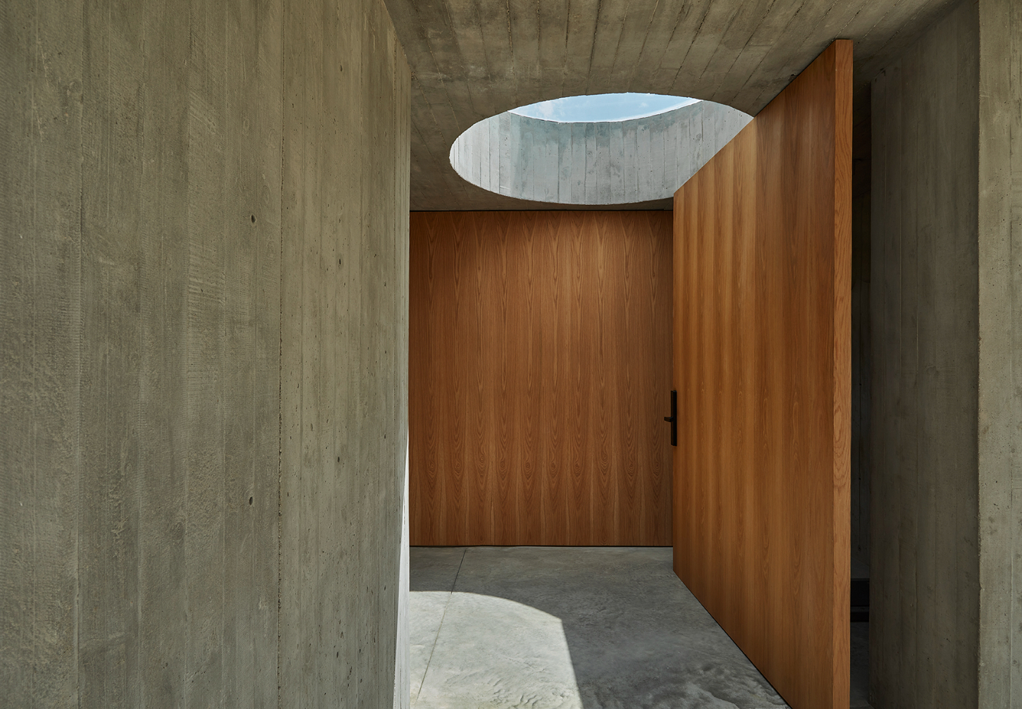 Casa Haras by 5Solidos — A concrete passage opens into a timber-lined interior, framed by board-formed concrete walls that establish a stark geometric threshold.