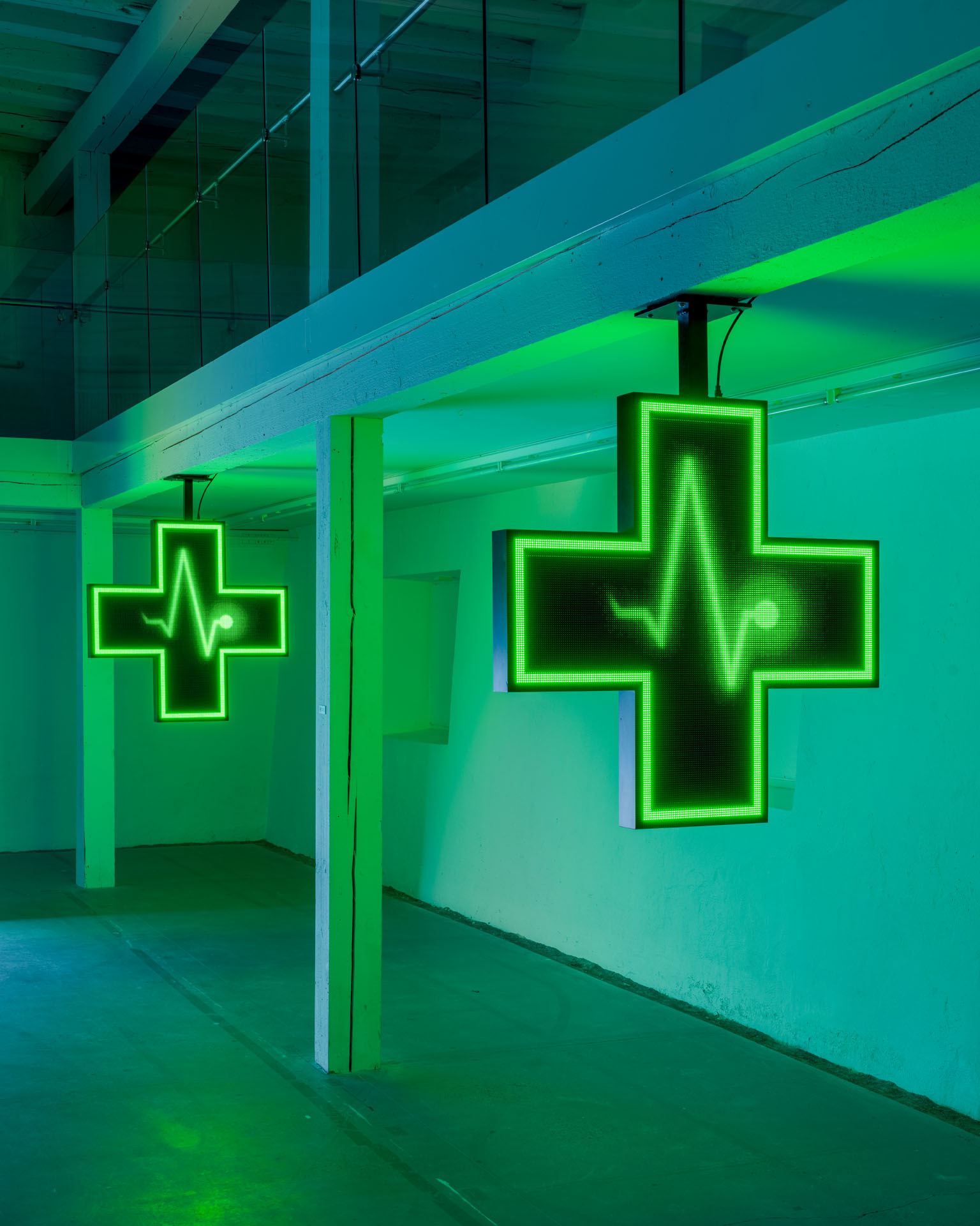 Milk Plus by Jens Settergren Viborg Kunsthal — Neon medical cross signs glow in acidic green against teal-lit concrete walls within an industrial gallery space.