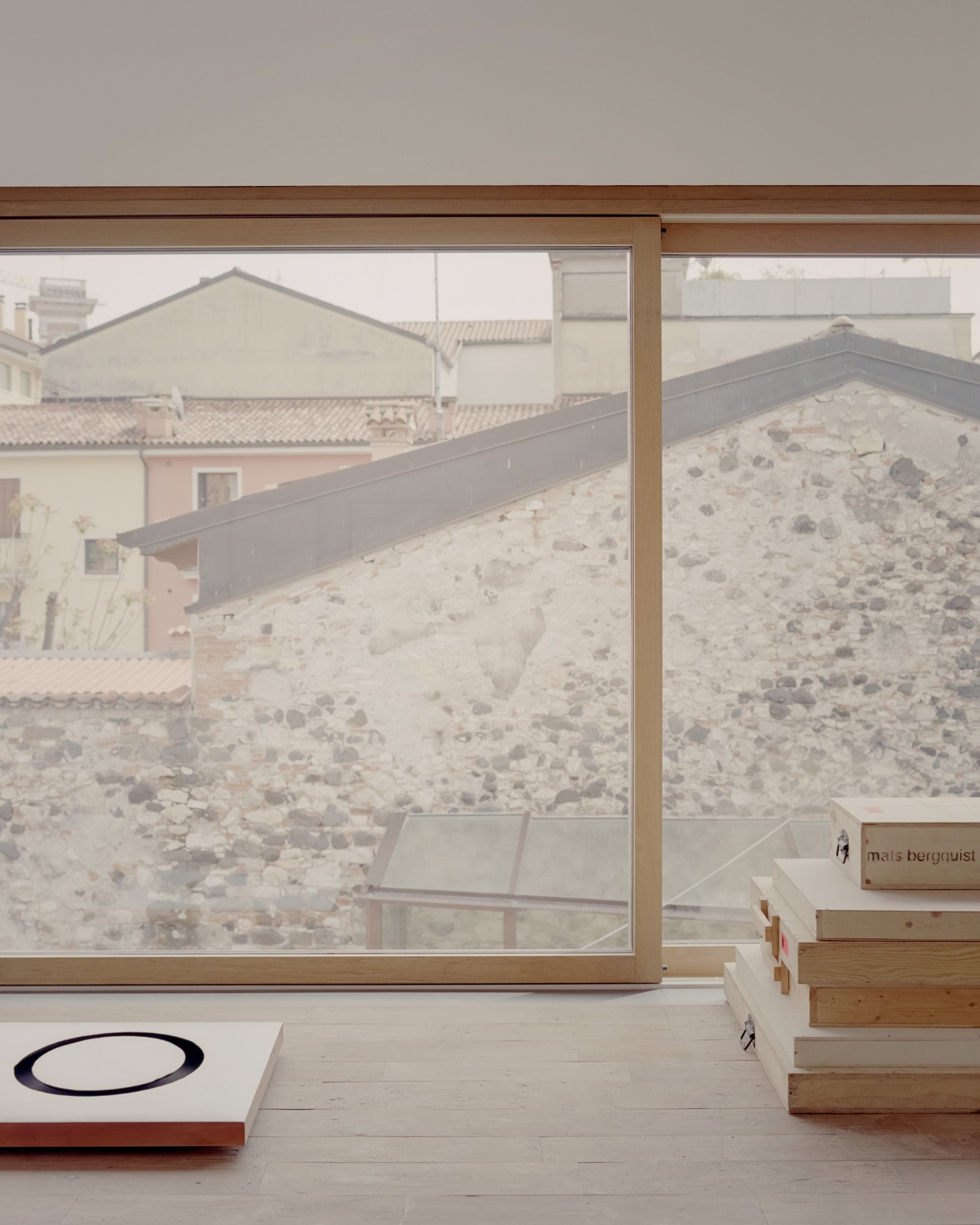 Atipografia Threshold and Treasure Gallery by AMAA — The threshold gallery frames a gravel forecourt and distant stone fortification through expansive glazing set in timber framing.