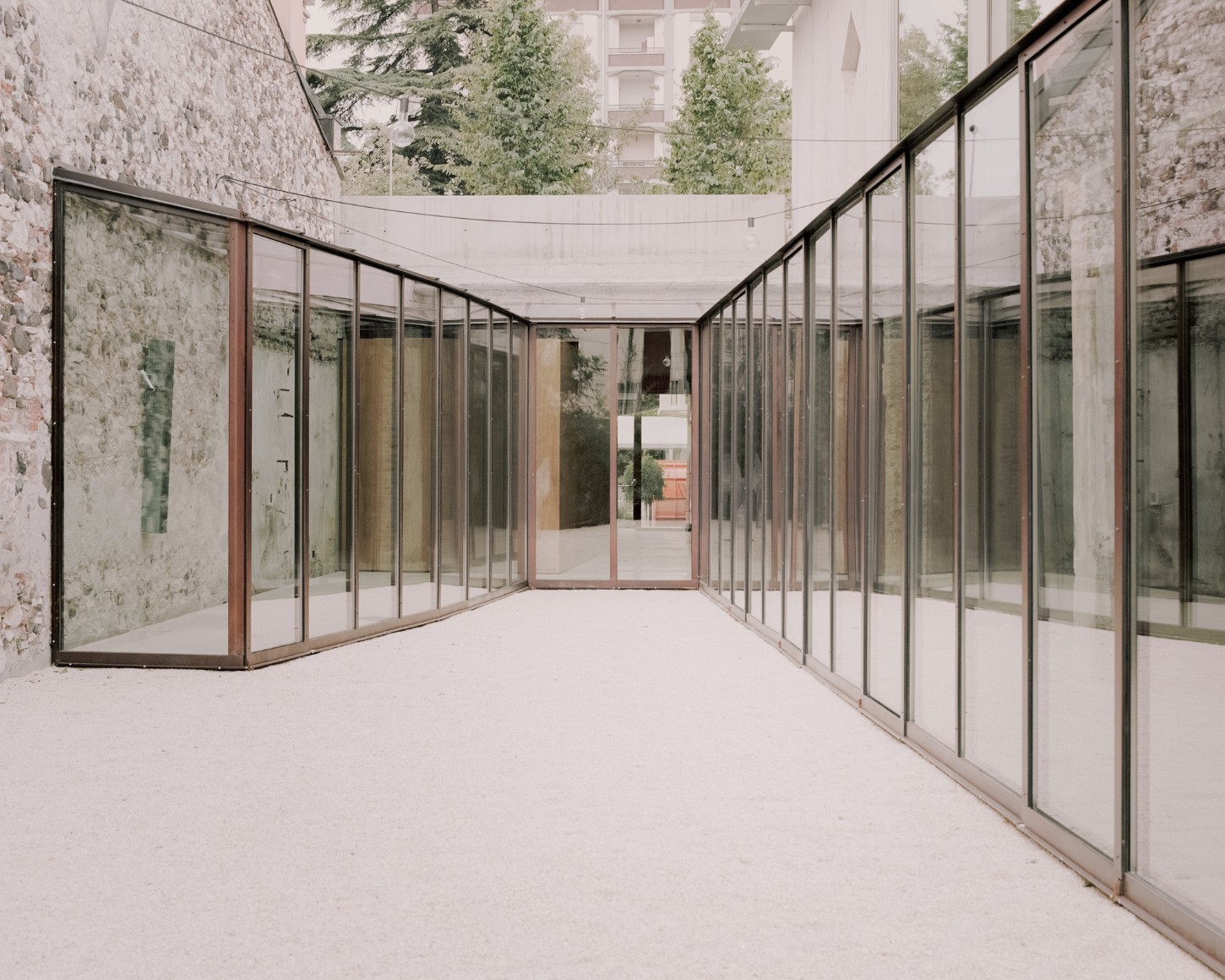 Atipografia Threshold and Treasure Gallery by AMAA — A glazed pavilion with metal-framed glass walls and doors extends into a gravel forecourt, flanked by weathered stone walls.