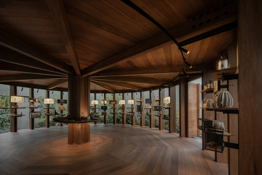 Xinú Marsella by Esrawe Studio by Estrawe Studio — A timber pavilion with radial shelving extends from a central circular wooden core, its cantilevered arms displaying ceramic vessels, glass bottles, and...