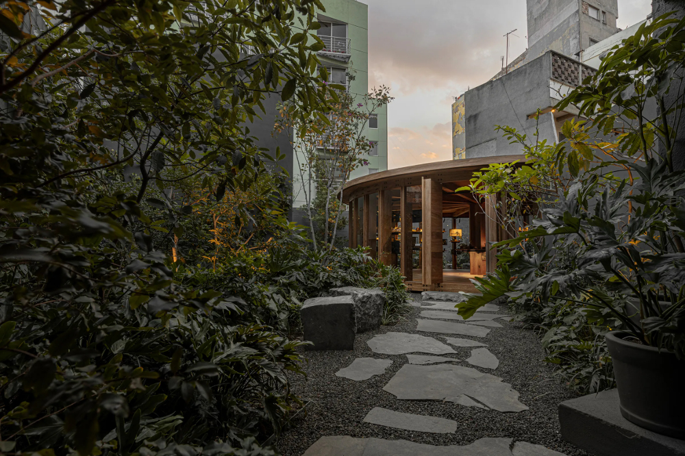 Xinú Marsella by Esrawe Studio by Estrawe Studio — A curved timber pavilion emerges from dense vegetation, its rounded wooden structure housing shelving and retail displays.