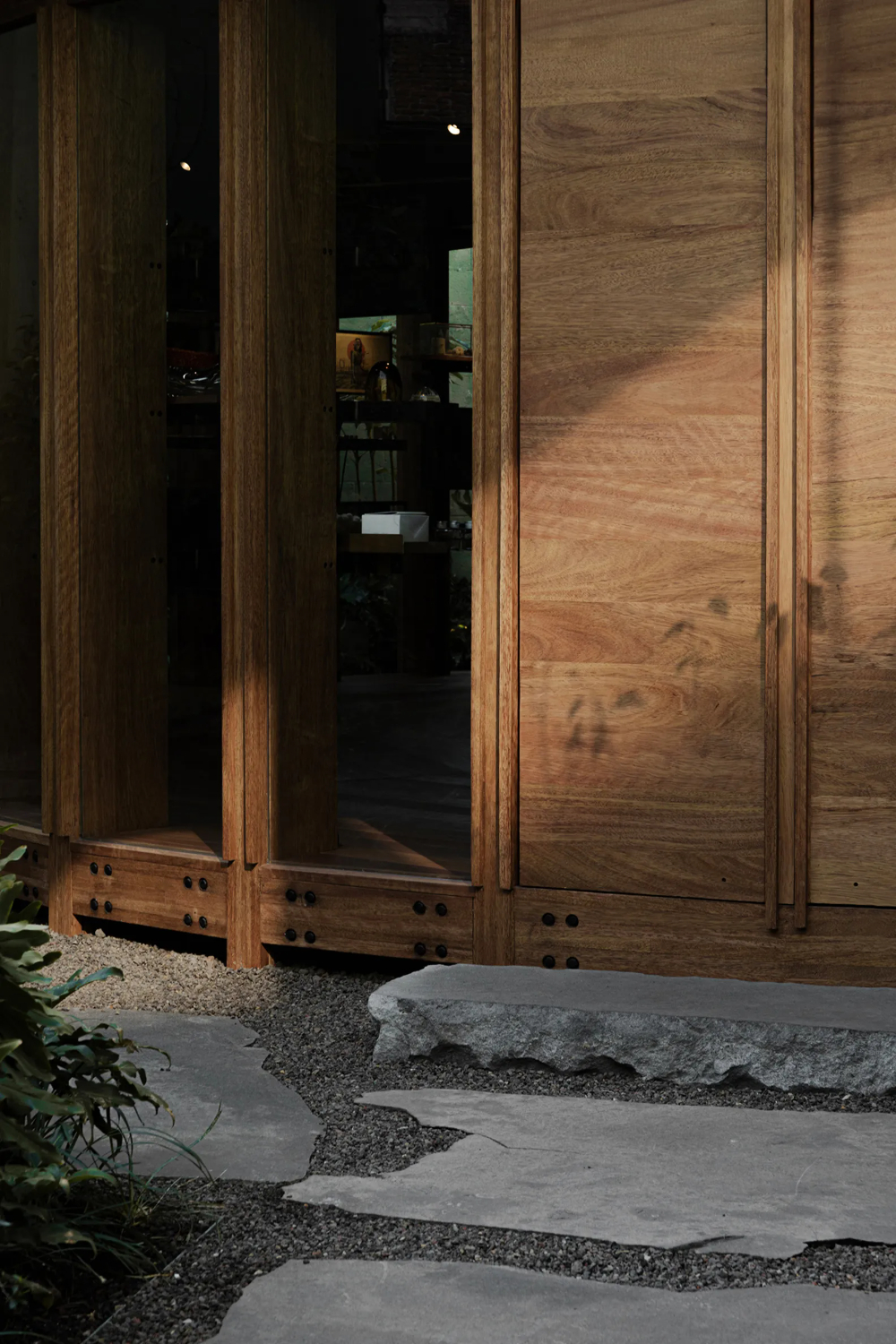 Xinú Marsella by Esrawe Studio by Estrawe Studio — Timber-framed glazed doors open onto a garden threshold, their warm wood surfaces casting long shadows across the gravel and stone stepping pathway.