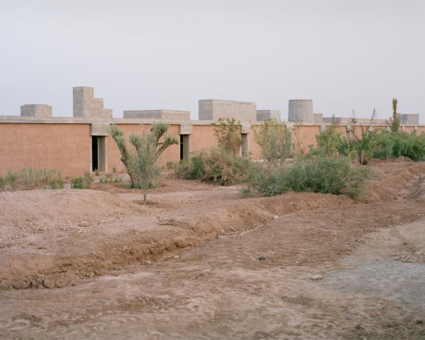 Dar El Farina by Leopold Banchini — Dar El Farina extends horizontally across an arid landscape, its terracotta-rendered walls punctuated by dark rectangular openings that establish a rhythmic...