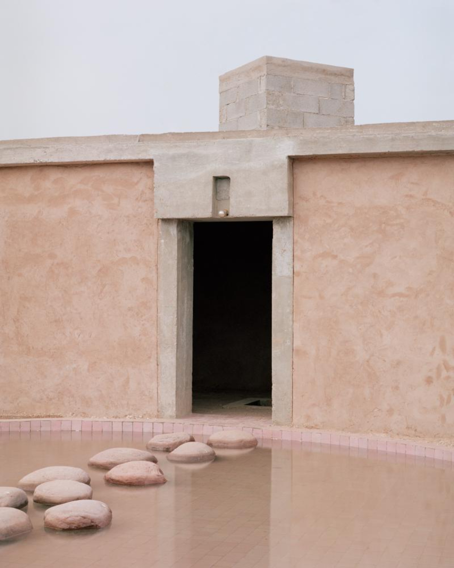 Dar El Farina by Leopold Banchini — A rendered plaster facade in terracotta and cream tones frames a deep recessed entrance, its geometric severity softened by organic stone elements scattered...