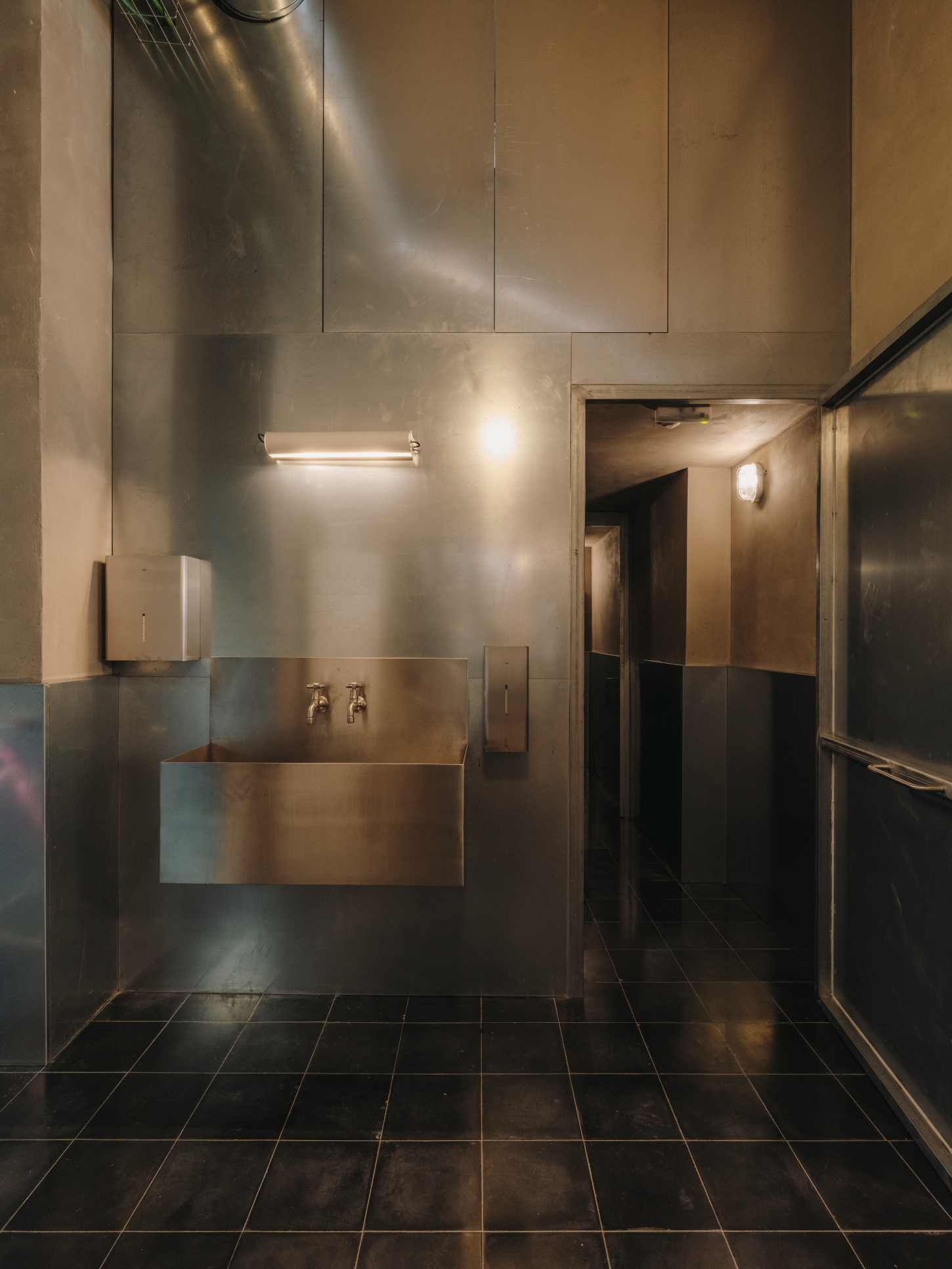 Parking Pizza by Skye Maunsell + Jordi Veciana — A commercial restroom designed with industrial minimalism, featuring stainless steel wall panels that create a clinical, hygienic environment.