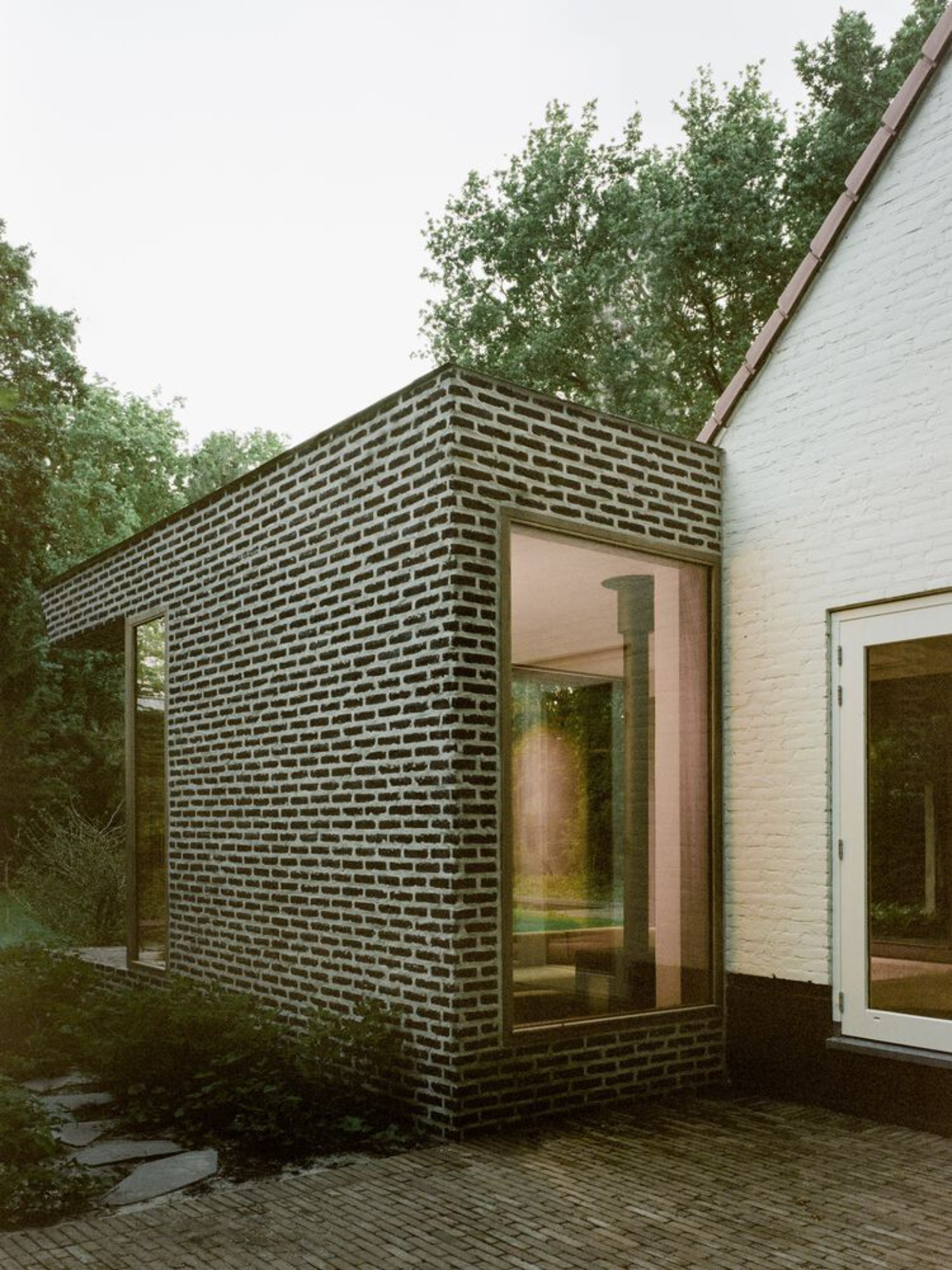 BD House by Space Encounters + Studio Vincent Architecture — A dark brick extension projects from the original white-rendered house, its facade defined by tight horizontal coursing that creates pronounced striping...