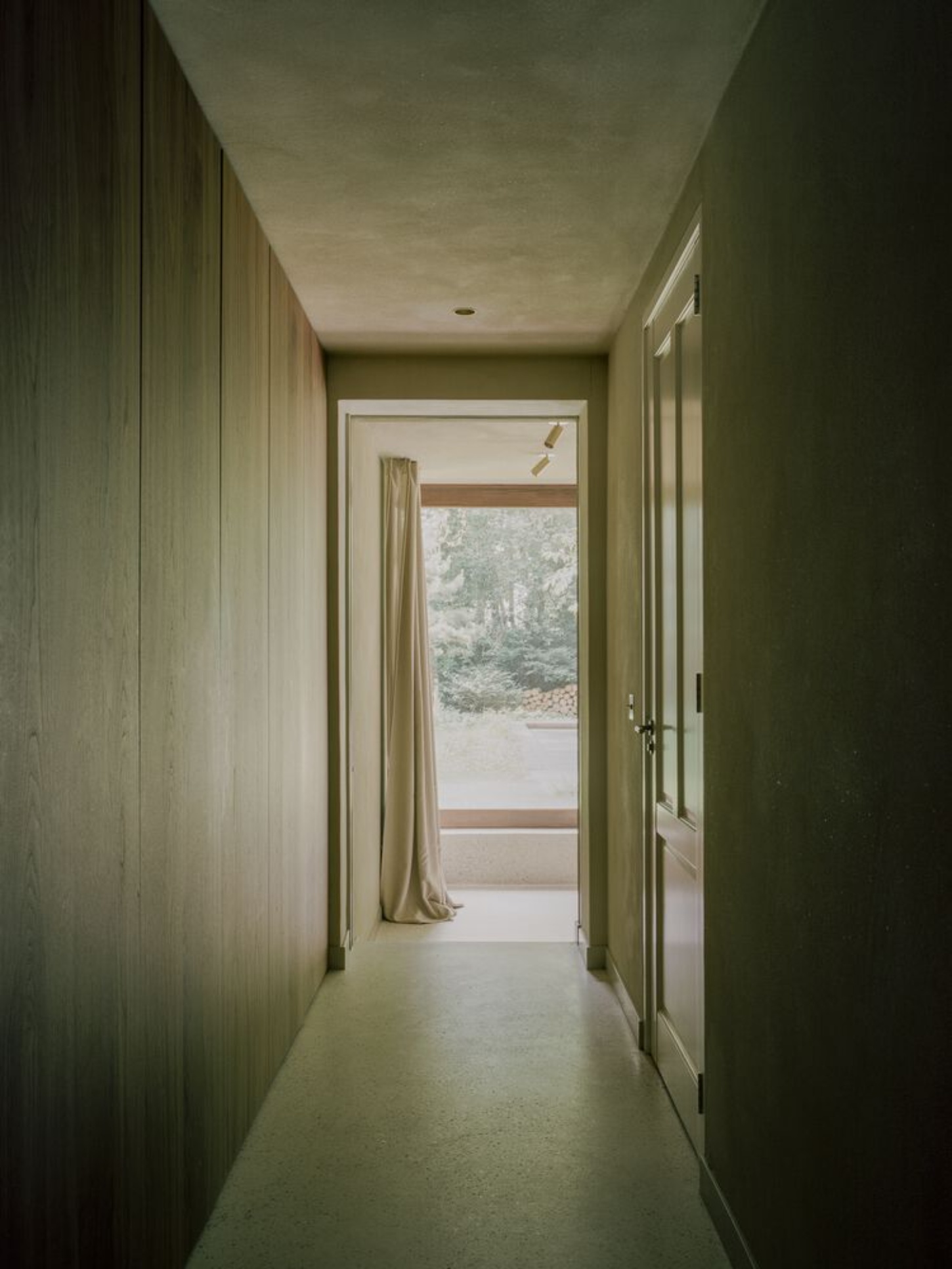 BD House by Space Encounters + Studio Vincent Architecture — A corridor extends toward glass doors framing the exterior landscape, its length emphasized by vertical textured walls in soft green-grey tones.