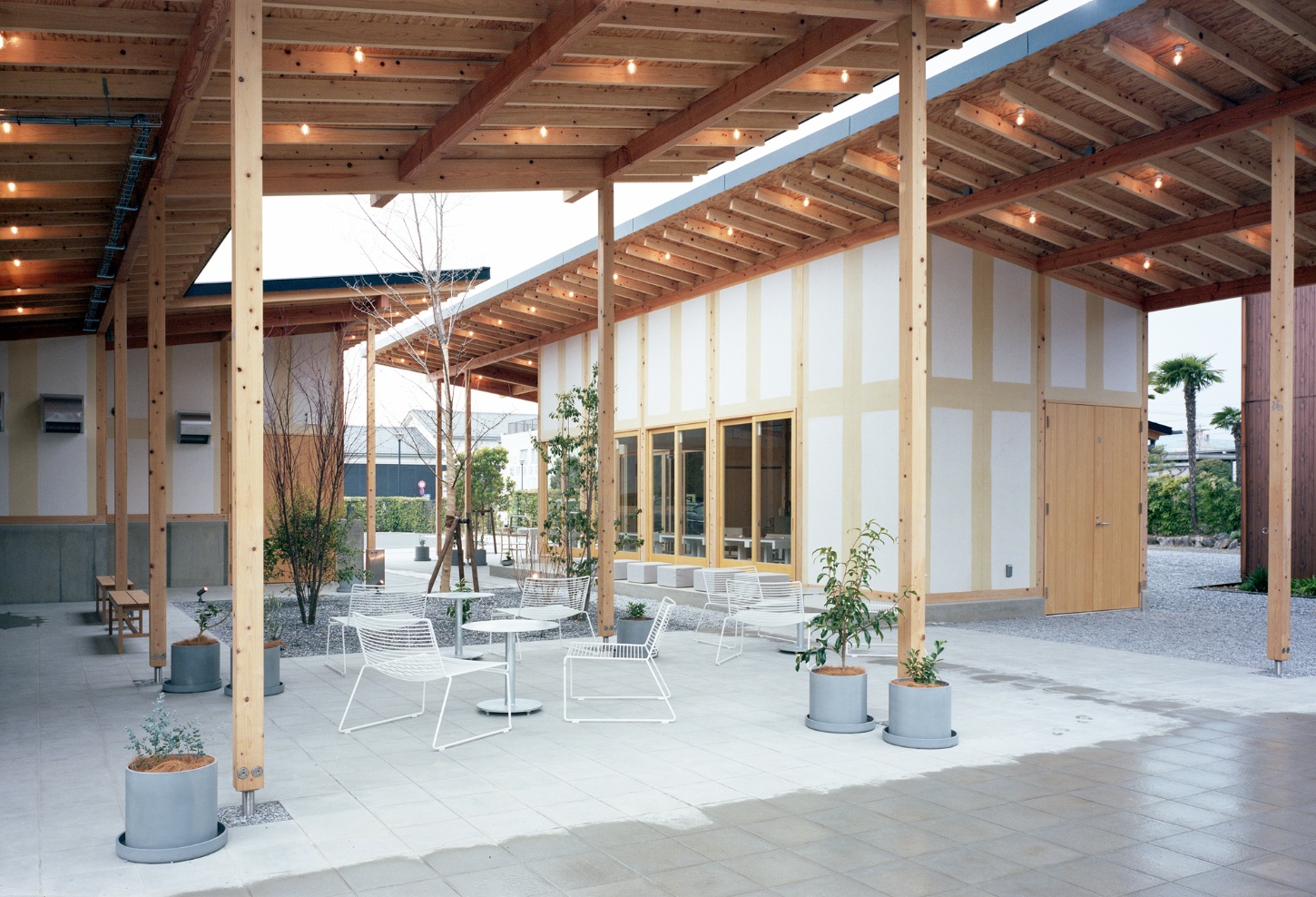 Funamachi Base Store by Jo Nagasaka + Schemata Architects — Beneath a post-and-beam timber pergola, a continuous concrete courtyard accommodates white powder-coated wire lounge chairs arranged in conversational clusters.