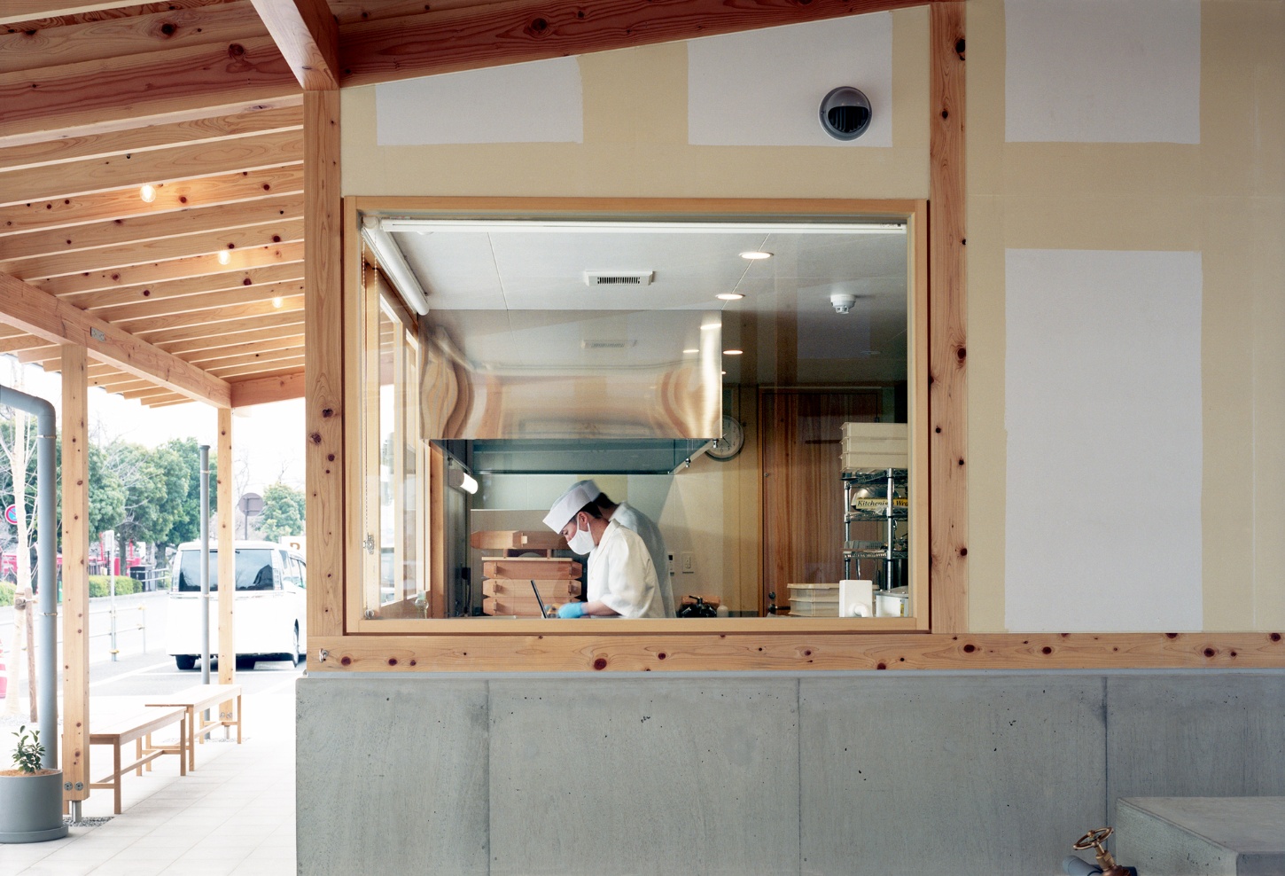 Funamachi Base Store by Jo Nagasaka + Schemata Architects — A service counter frames a worker in white protective gear and mask, visible through a large timber-framed glazed opening.