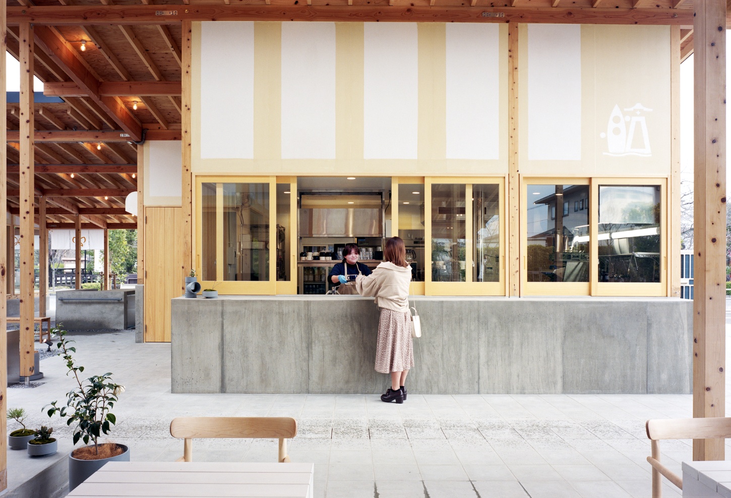Funamachi Base Store by Jo Nagasaka + Schemata Architects — A customer stands at a concrete service counter beneath a timber post-and-beam structure, its regular grid of exposed fasteners visible overhead.