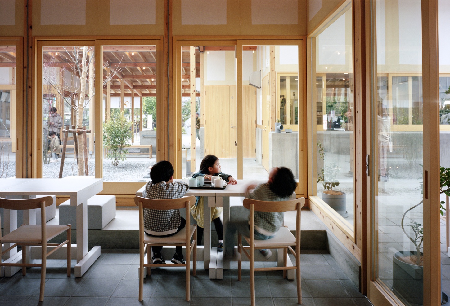 Funamachi Base Store by Jo Nagasaka + Schemata Architects — Three figures occupy a timber-framed interior, seated at a white table positioned against continuous glazed doors that open to a courtyard landscape.
