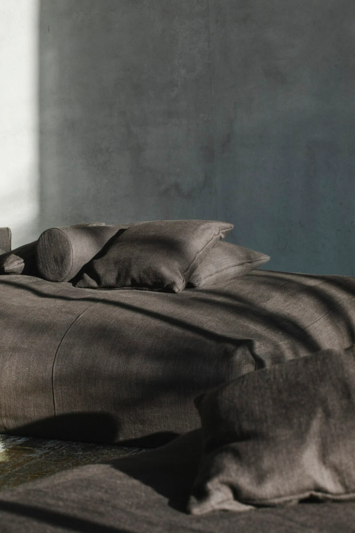 Villa Mahler by Anna Philipp + Eichkorn — Folded linen bedding in warm grey tones arranged on a polished concrete floor against a textured plaster wall.