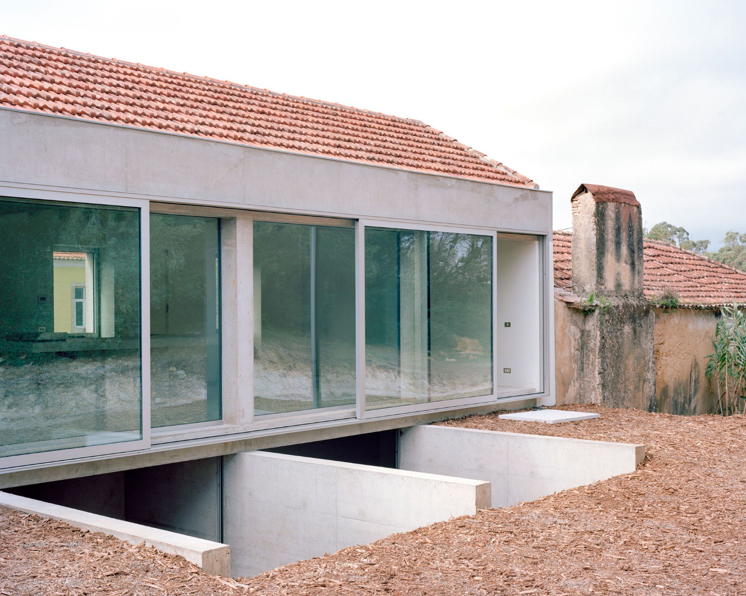 Tenant House by Arquitectura-G — A sunken concrete threshold frames the building's ground floor, its monolithic edges creating a stark geometric interruption in the ochre earth.