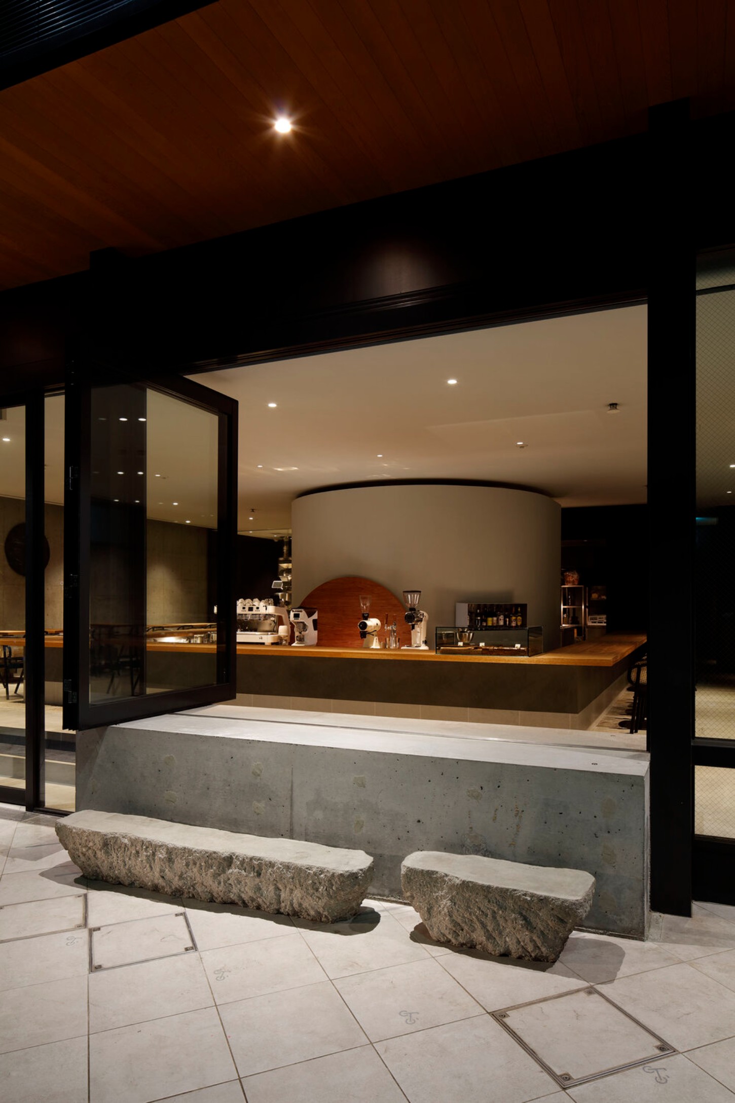 OGAWA COFFEE LABORATORY by Yusuke Seki Studio — The KURA bean cellar anchors this coffee laboratory interior as a monolithic cylindrical form rendered in white plaster, positioned centrally behind a...