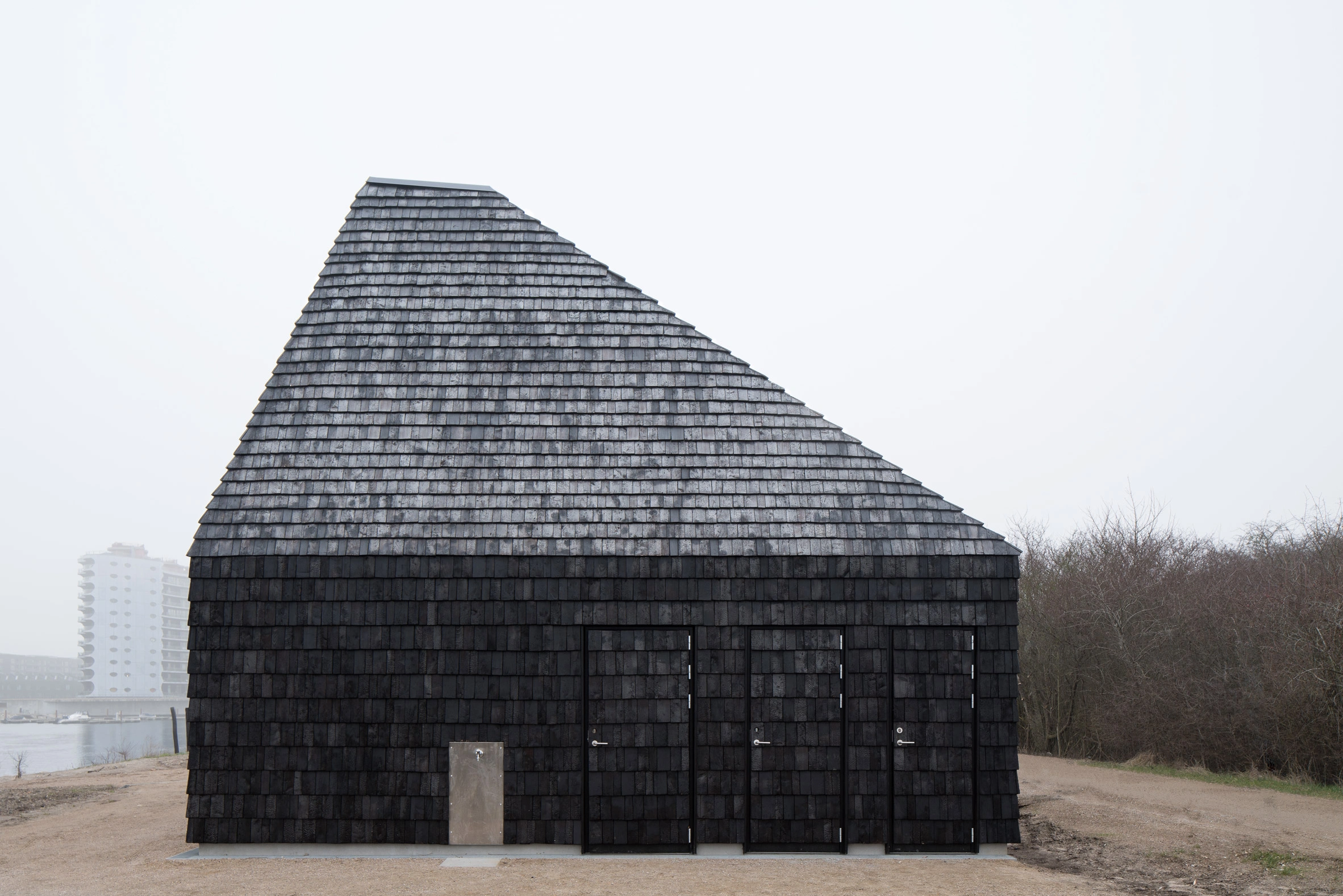 Amager Nature Park by ADEPT and LYTT Architecture — A visitor pavilion at Amager Nature Park presents a conical timber structure clad entirely in dark cedar shingles, its pitched roof tapering sharply to an apex.
