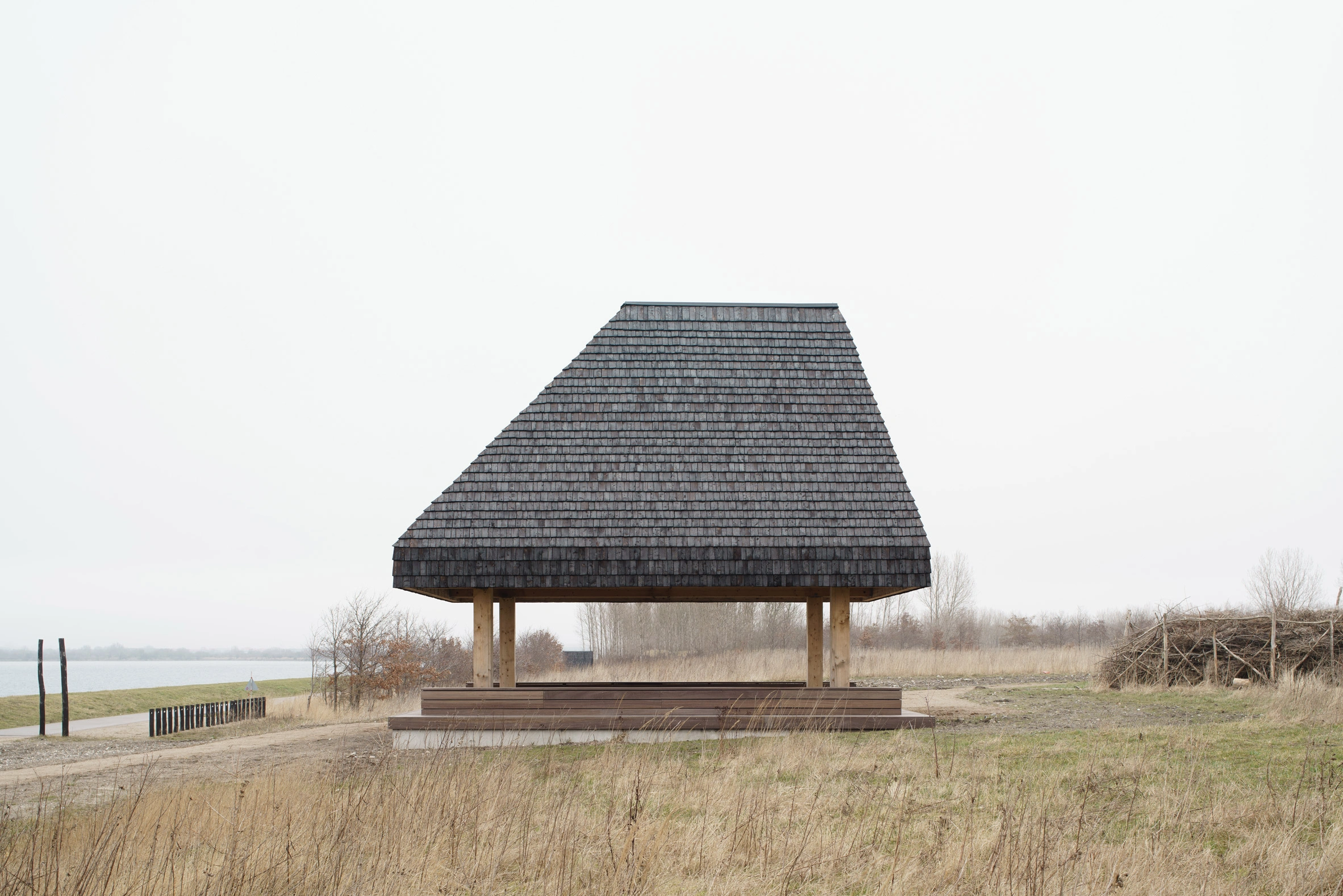 Amager Nature Park by ADEPT and LYTT Architecture — A pavilion structure dominates the winter landscape of Amager Nature Park, its conical roof clad in dark cedar shingles tapering to a sharp apex.