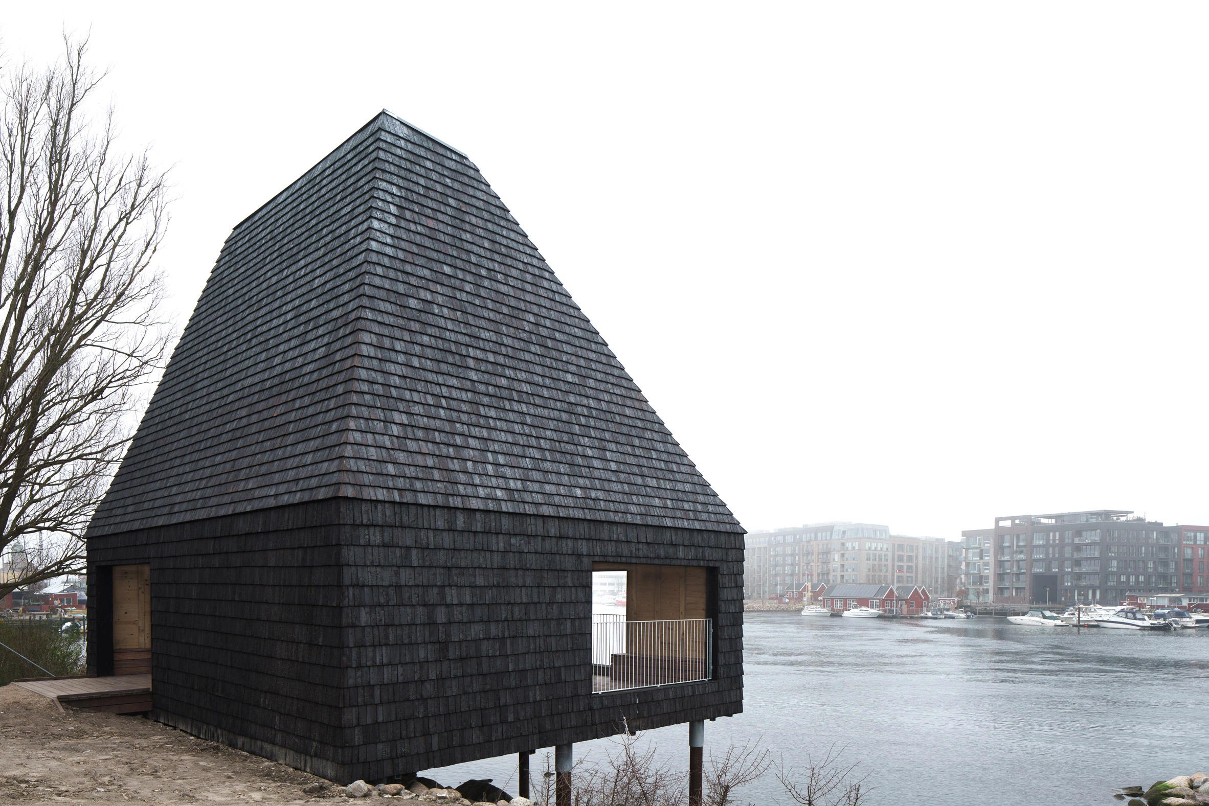 Amager Nature Park by ADEPT and LYTT Architecture — A conical timber-clad pavilion anchors the water's edge at Amager Nature Park, its peaked roof tapering sharply to an apex above the flat Copenhagen waterscape.