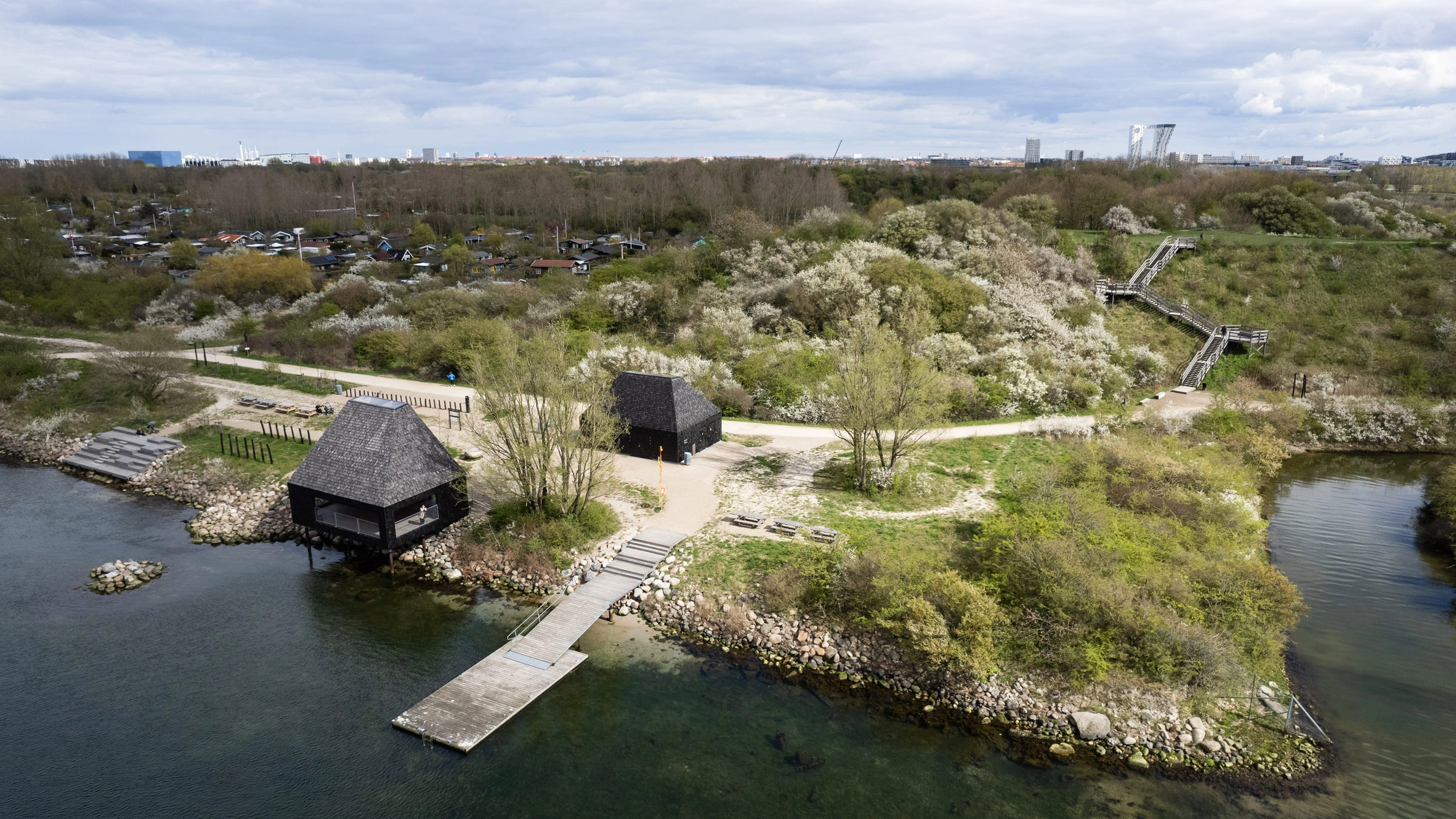 Amager Nature Park by ADEPT and LYTT Architecture — Aerial view of Amager Nature Park reveals two dark-clad pavilions positioned on wooden pilings above water and rocky substrate.