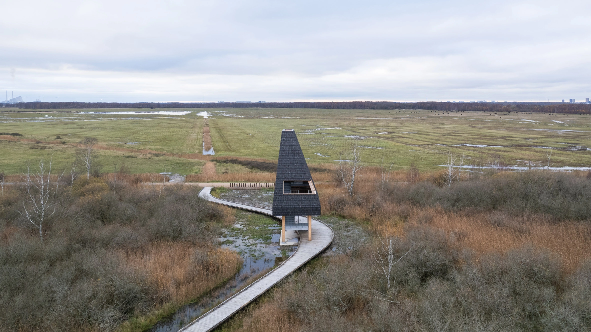 Amager Nature Park by ADEPT and LYTT Architecture — A conical timber-clad pavilion anchors the wetland landscape of Amager Nature Park, its dark cedar shingle exterior tapering to a sharp apex.