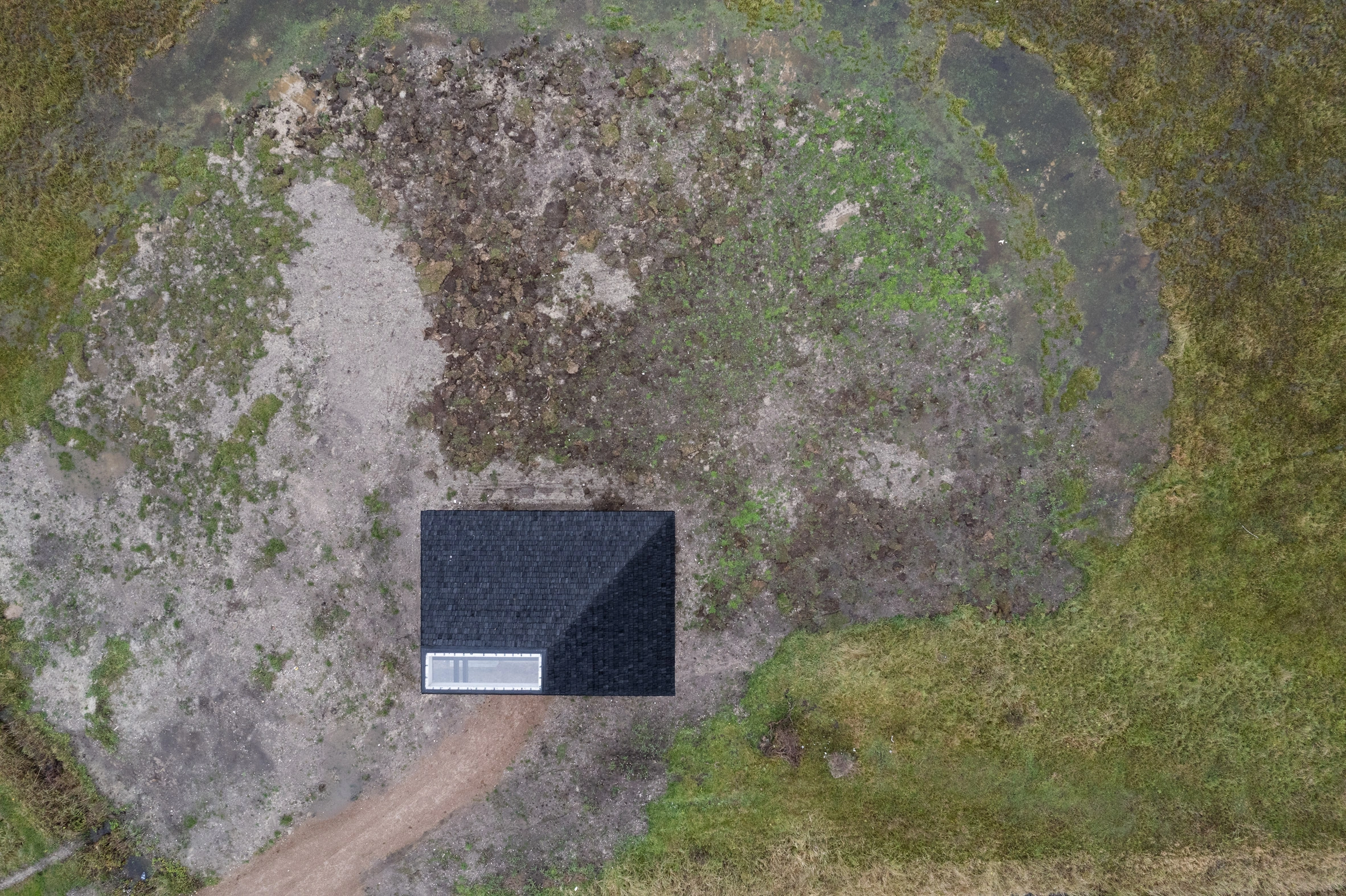 Amager Nature Park by ADEPT and LYTT Architecture — Aerial view of a visitor pavilion at Amager Nature Park reveals its minimalist dark-clad form set against moss-covered ground and surrounding vegetation.
